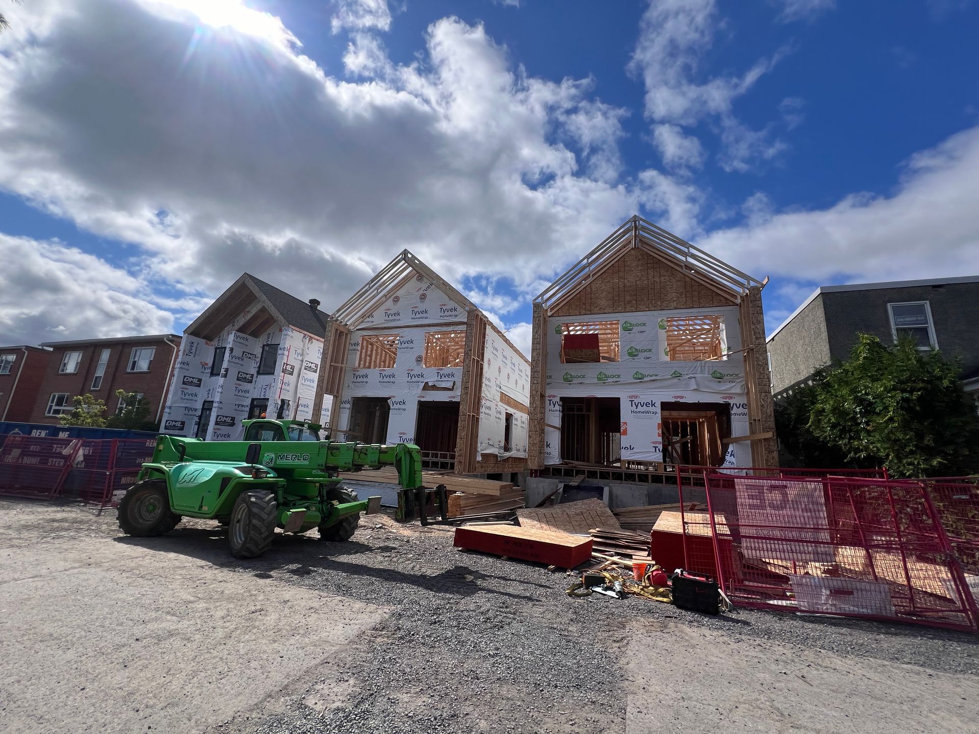 Trois maisons en construction, en cours de finition, derrière une clôture, avec un véhicule utilitaire vert devant, sous un ciel bleu nuageux.
