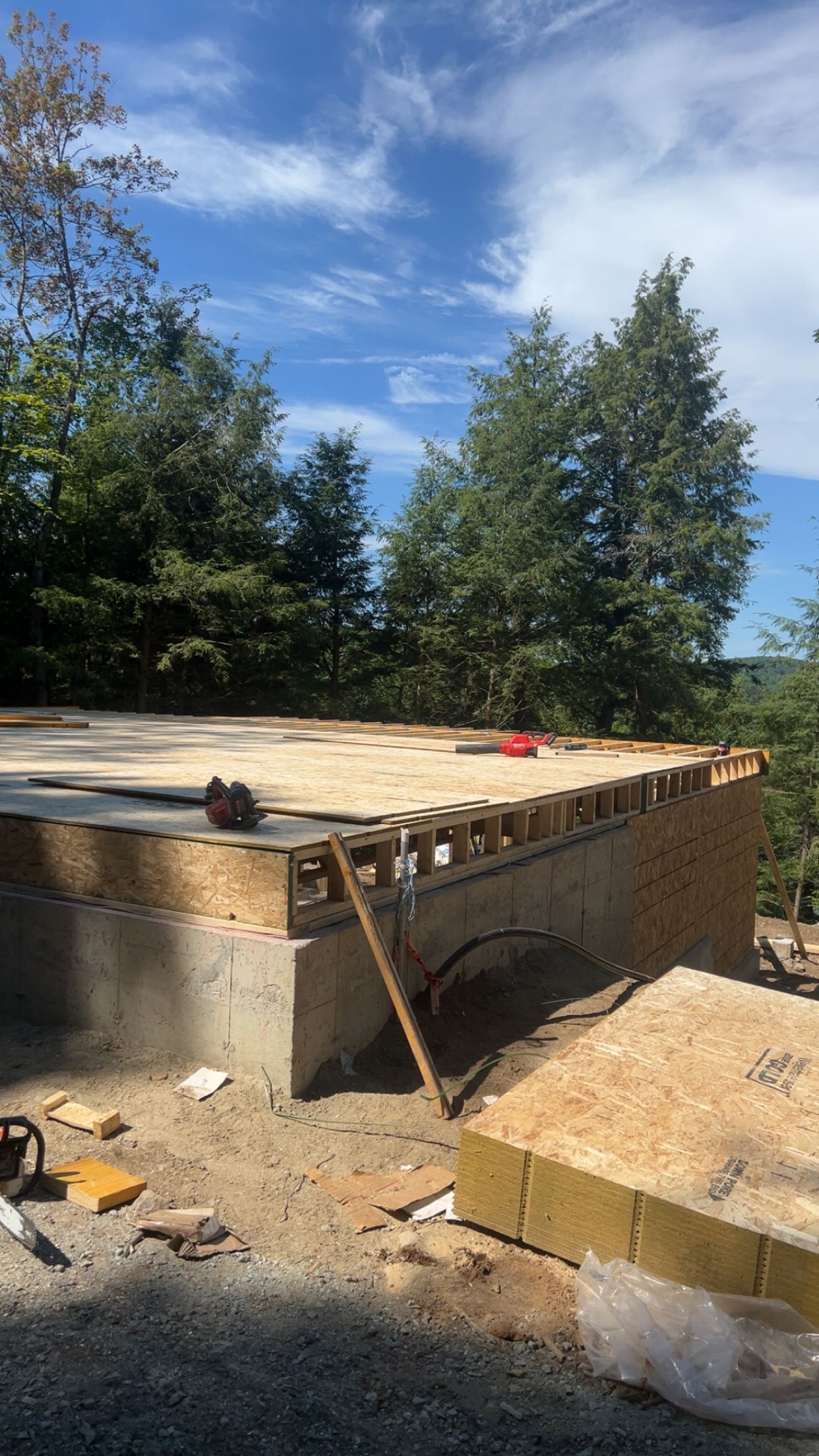 Dalle de fondation en béton en construction sur un terrain boisé, avec du bois, des outils et un ciel bleu au-dessus.