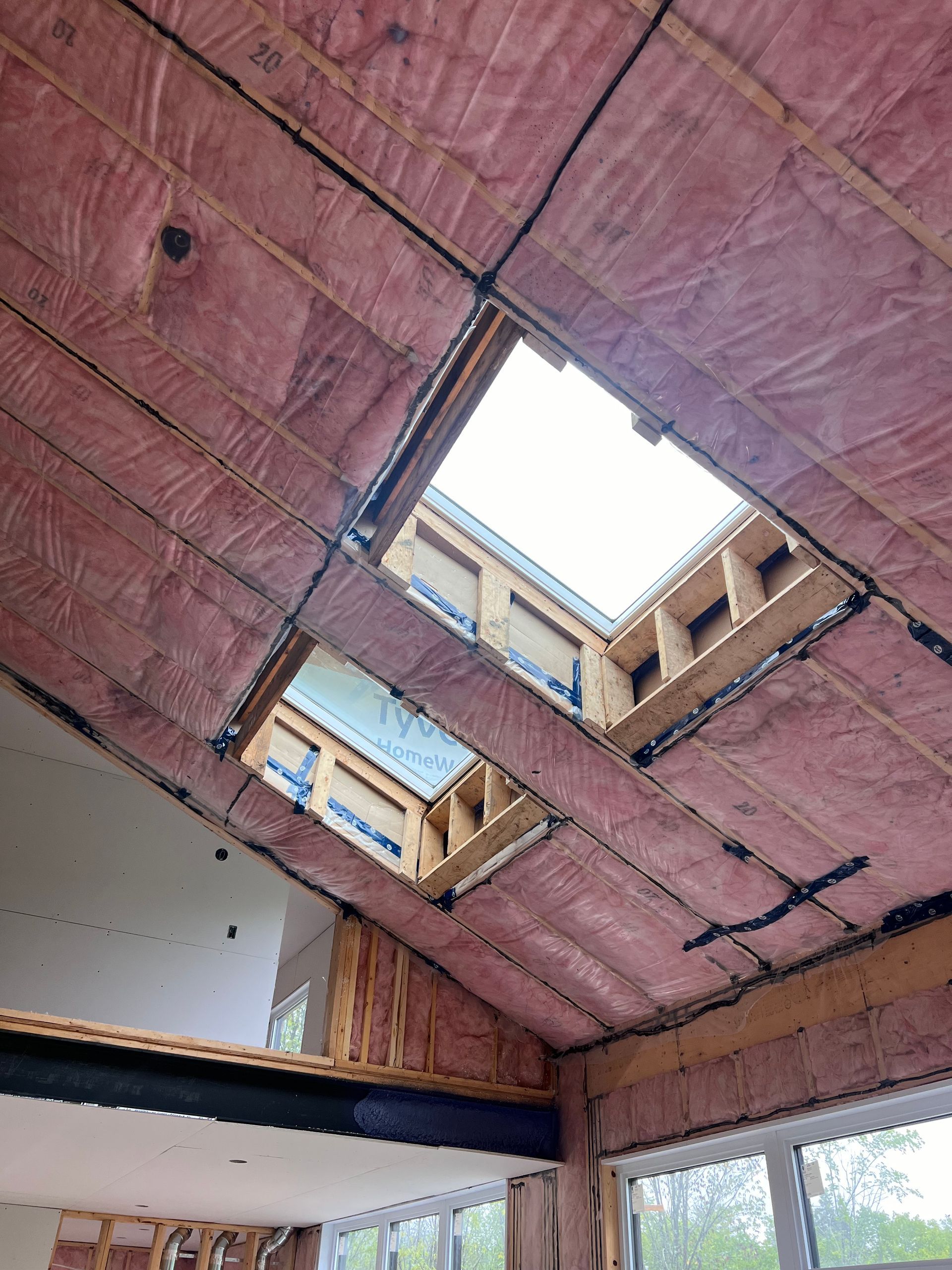 Ouverture au plafond d'un bâtiment en construction, avec isolation rose et poutres apparentes autour d'un puits de lumière.