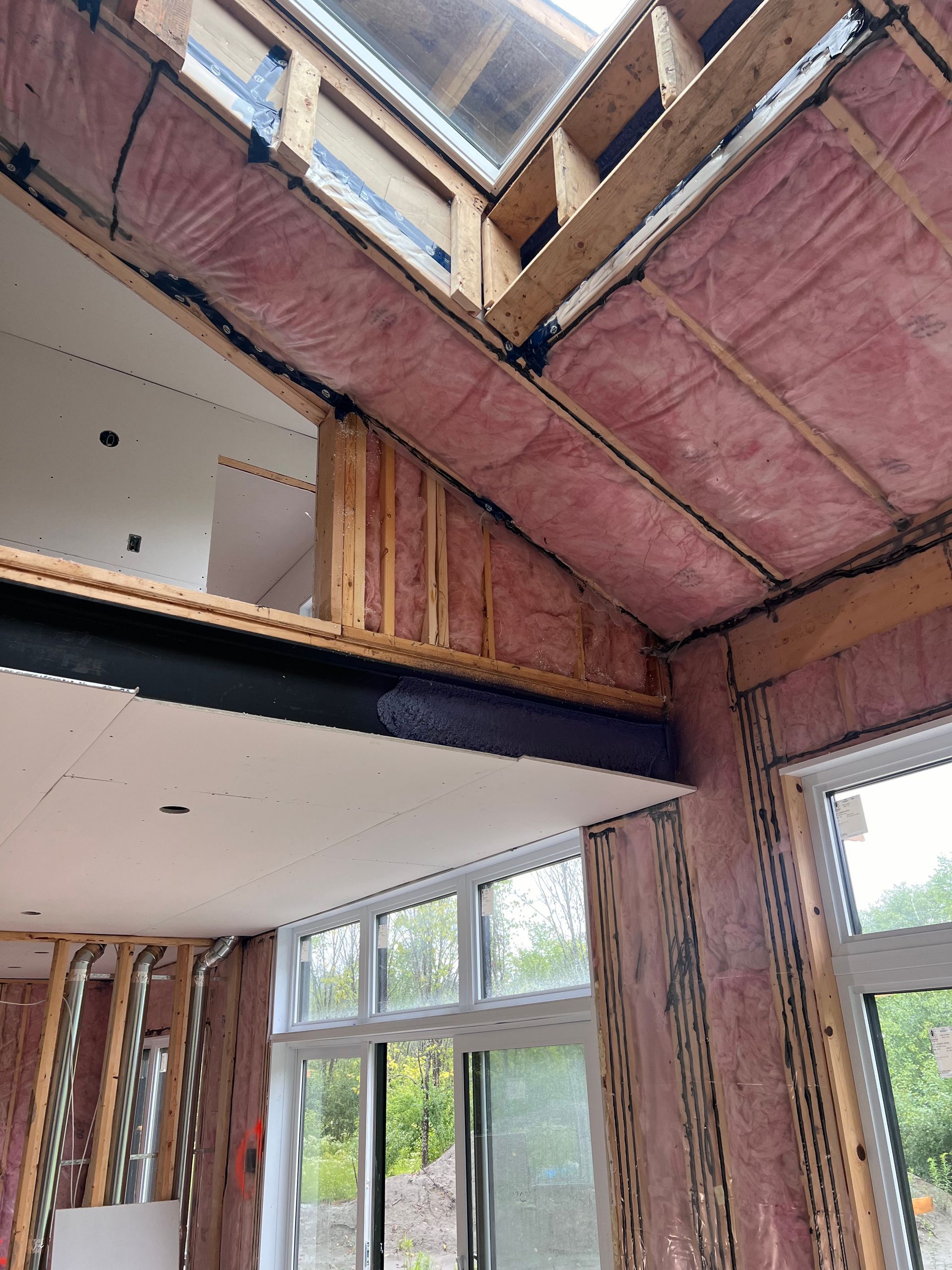 Charpente intérieure apparente et isolation rose dans une maison en construction, avec de grandes fenêtres et un puits de lumière.
