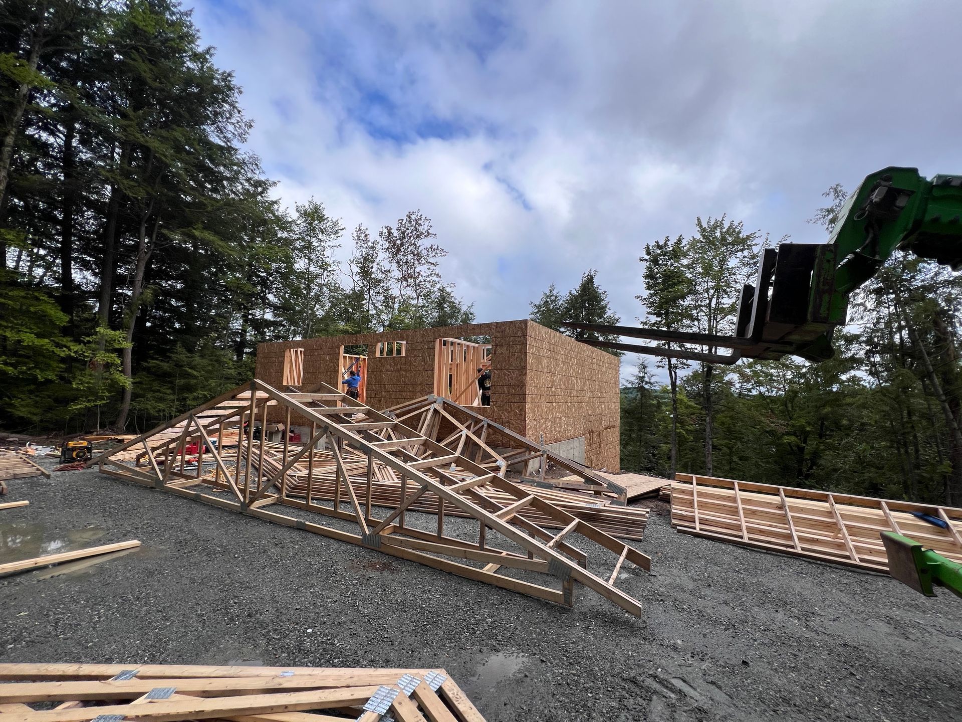 Maison à ossature bois en construction, avec des piles de bois et des ouvriers sur un terrain de gravier, entourée d'arbres.