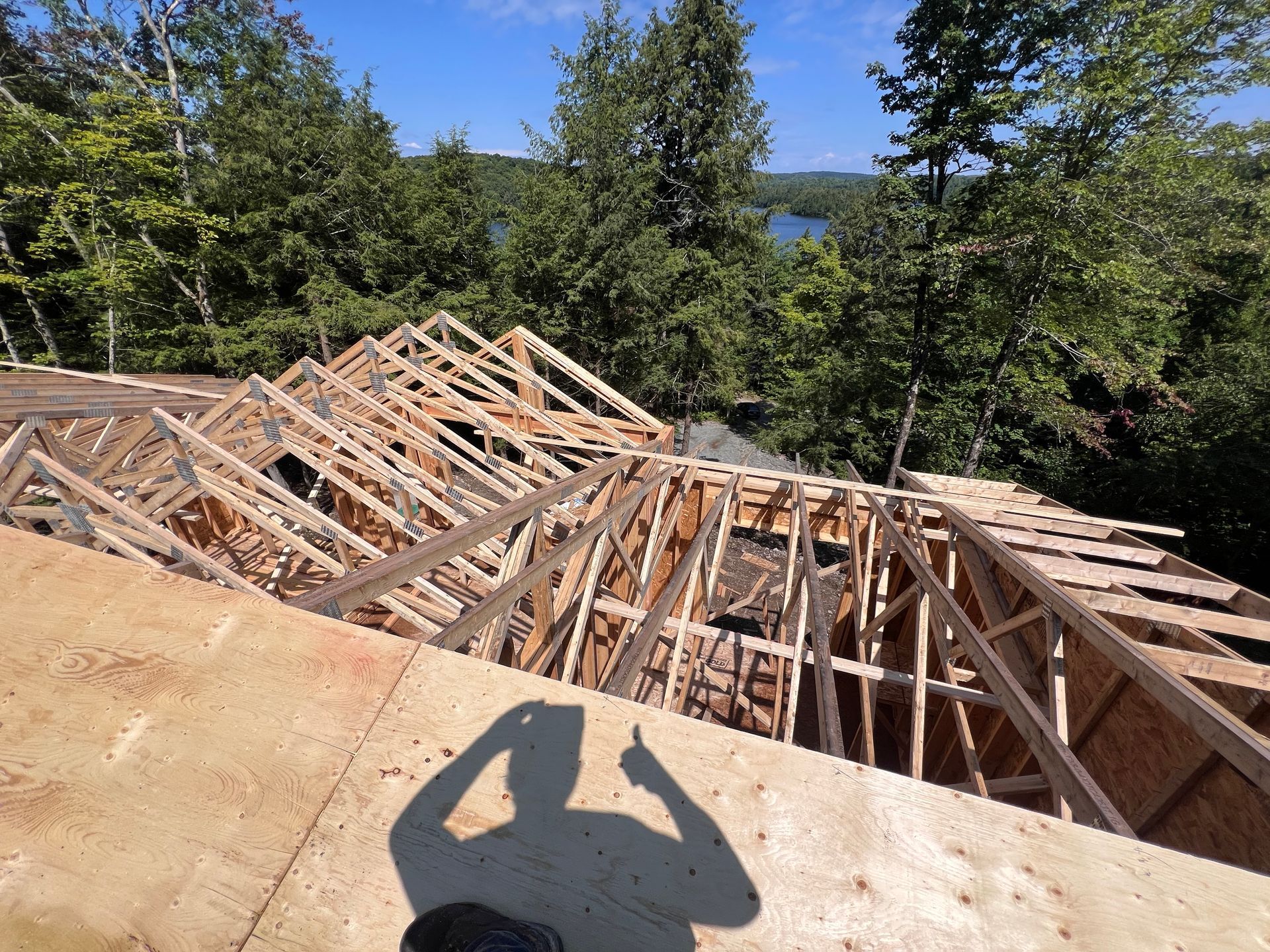 Charpente de toit en bois partiellement construite sur un terrain où se dresse une maison, avec des arbres et un lac en arrière-plan.