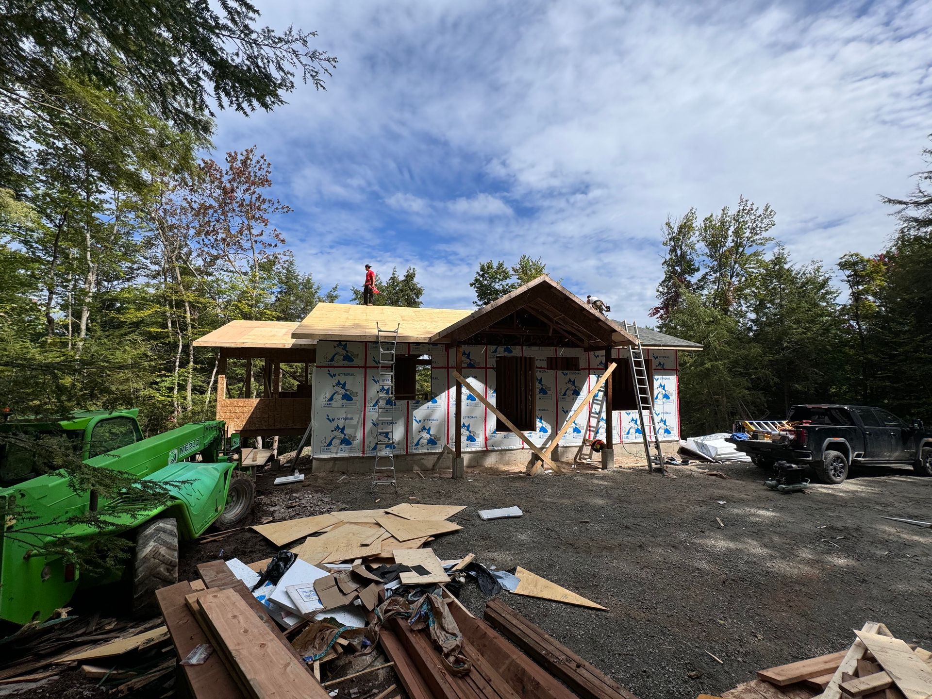 Cabane partiellement construite avec des ouvriers, un tracteur vert et un camion noir sur un terrain gravillonné entouré d'arbres