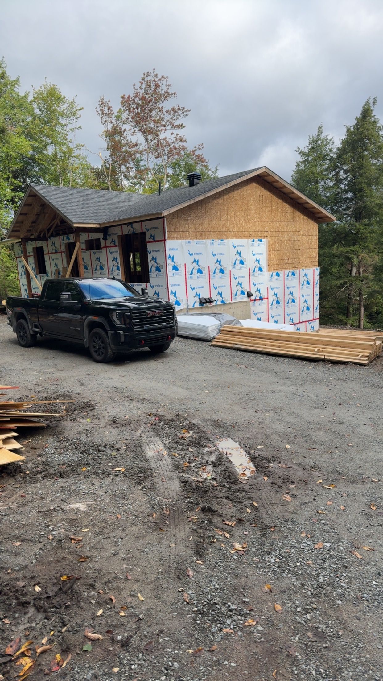 Cabane en construction, avec une camionnette noire garée sur du gravier, des murs recouverts de bâches et du bois empilé à proximité.