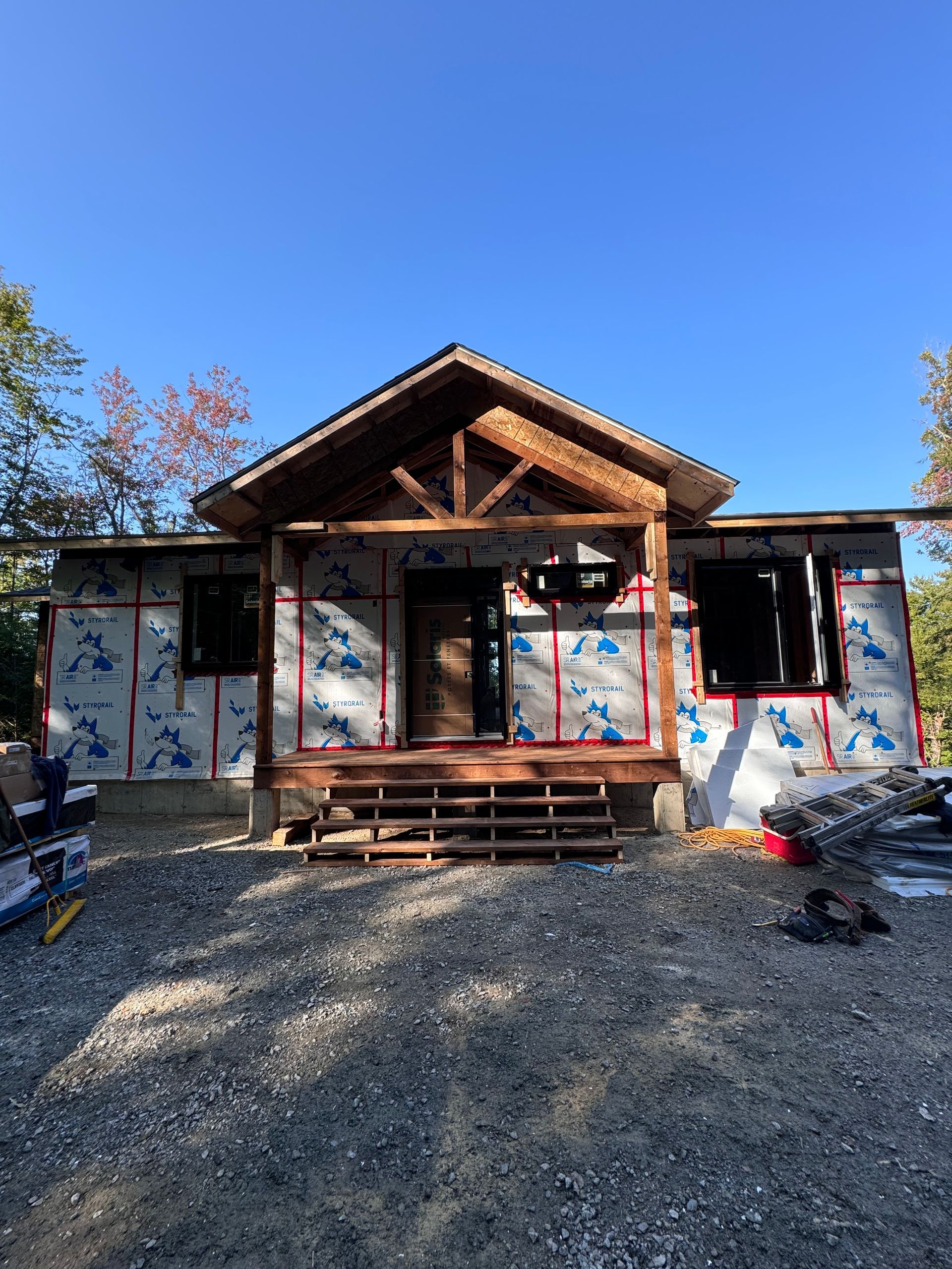 Petite maison en construction avec cour gravillonnée, ciel bleu et marches de porche en bois.