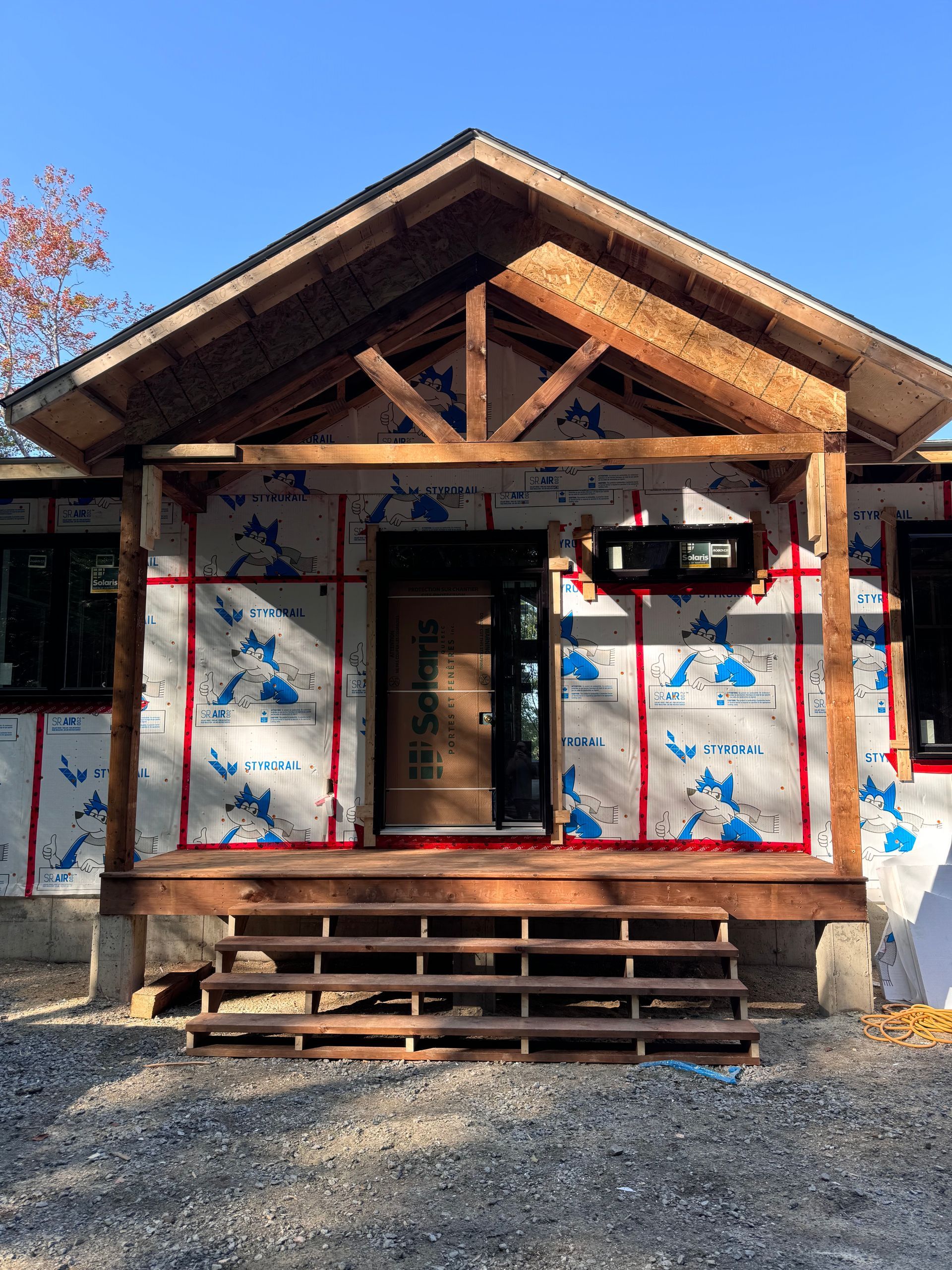 Petite maison en construction avec charpente apparente en bois, marches d'entrée et murs recouverts de revêtement, sur un terrain en gravier.