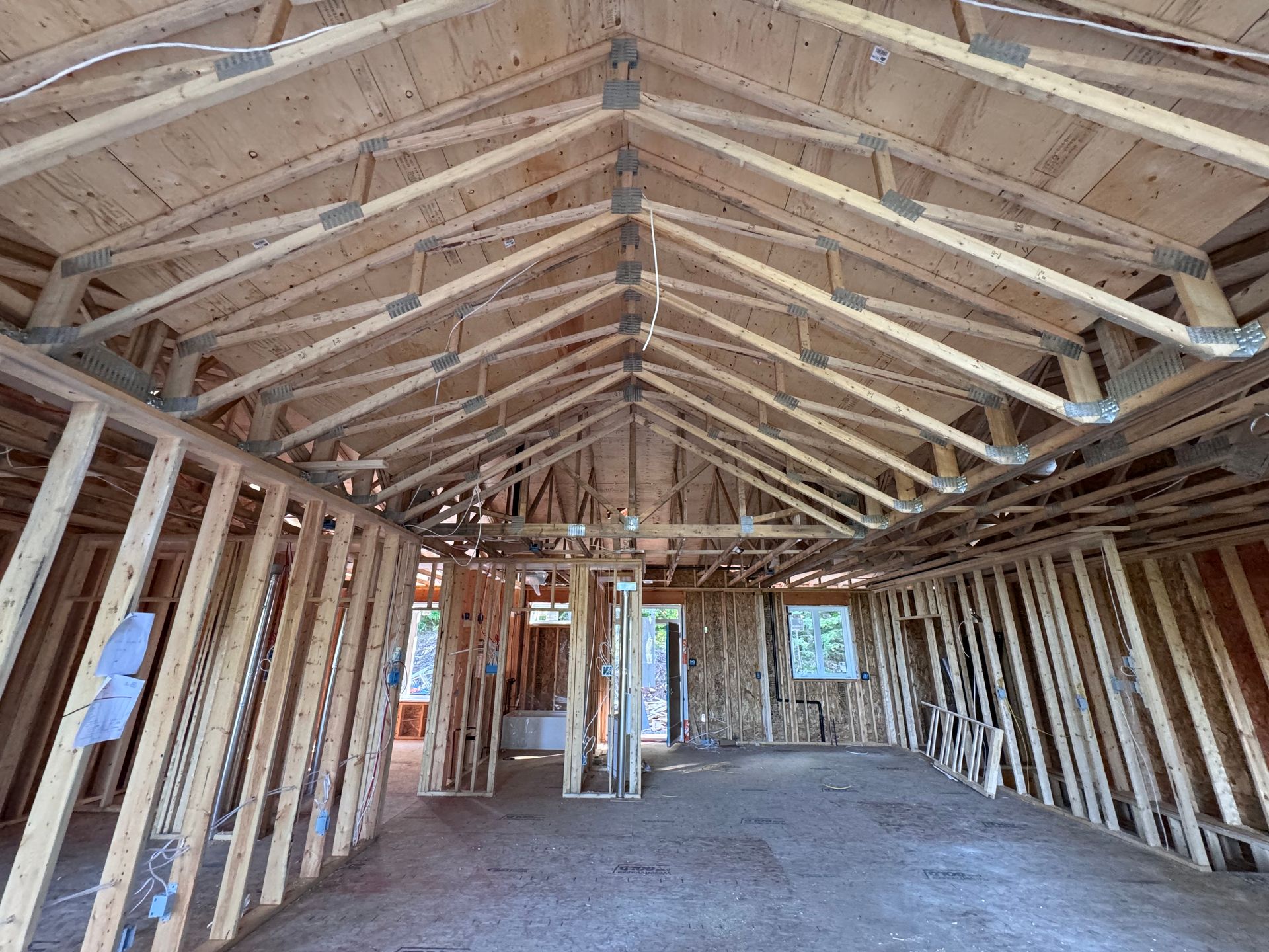 Intérieur d'une maison inachevée, avec charpente en bois apparente et fermes de toit, vu depuis une longue pièce ouverte.