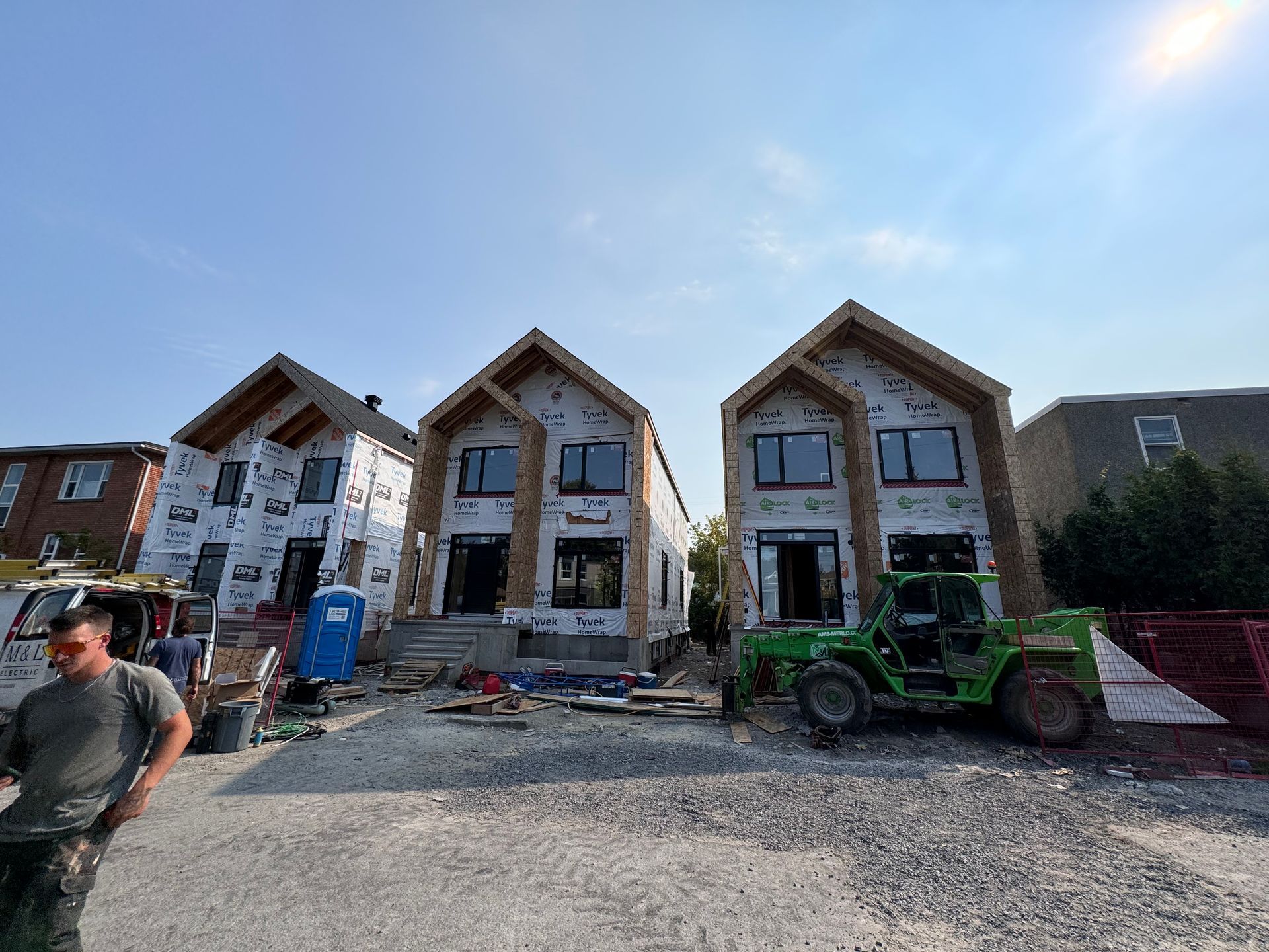 Trois façades de maisons de ville inachevées sur un chantier, avec un tracteur vert devant et des débris au sol.