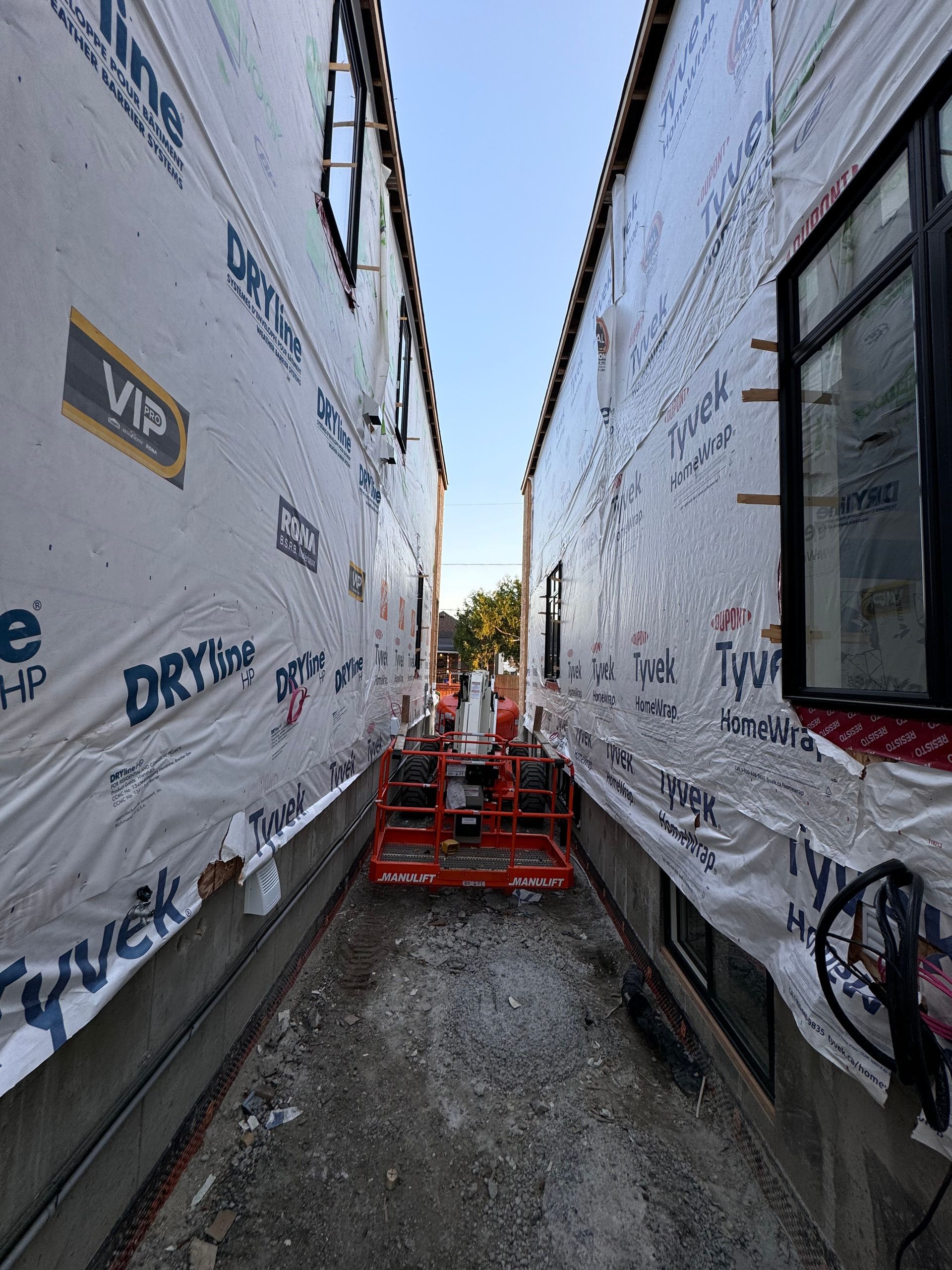 Ruelle étroite entre des murs recouverts de bâches, avec des échafaudages orange et un chemin de gravier en construction.