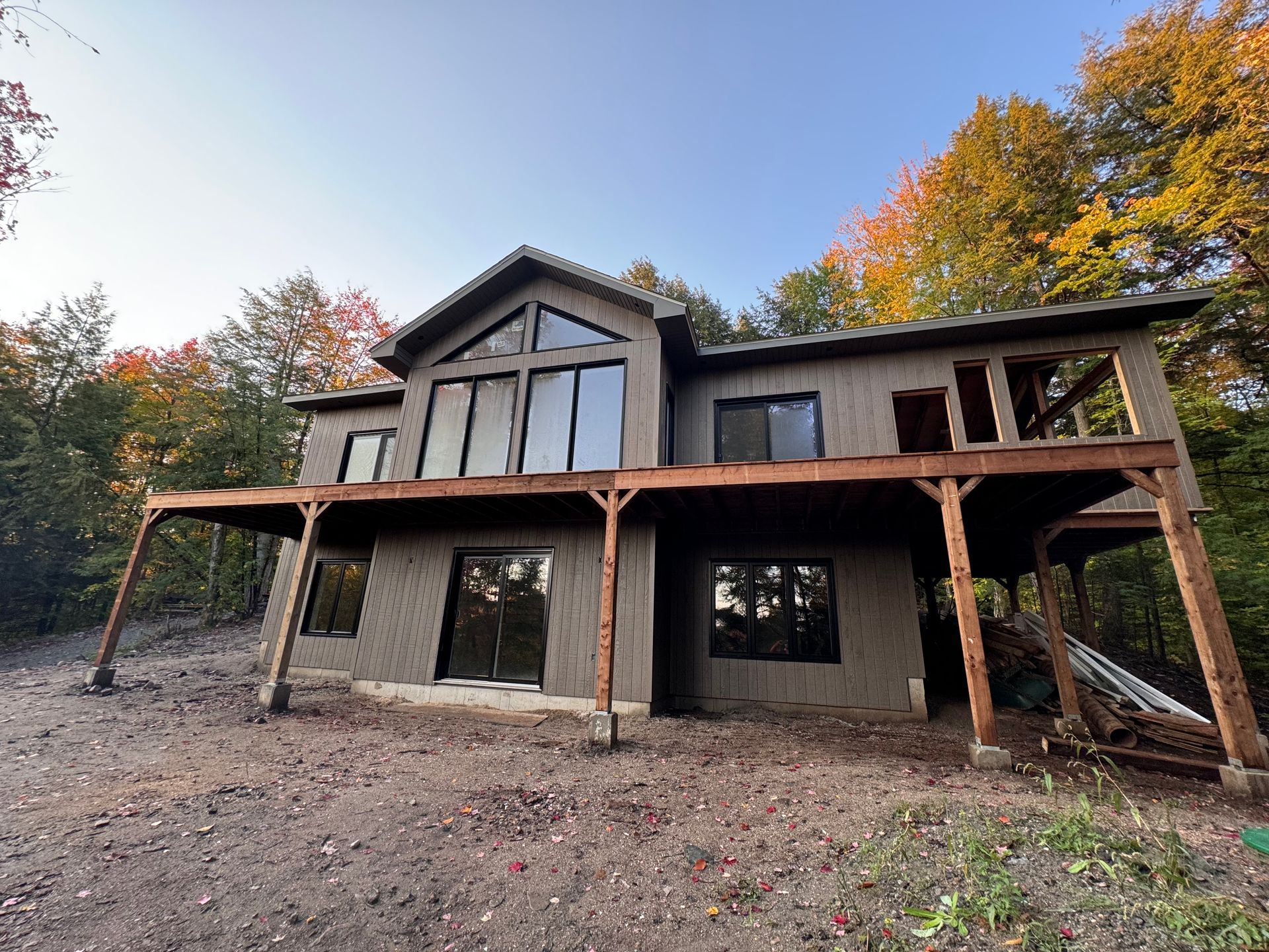 Maison moderne en construction dans une zone boisée, avec un extérieur en pierre et en bois et des arbres aux couleurs d'automne tout autour.