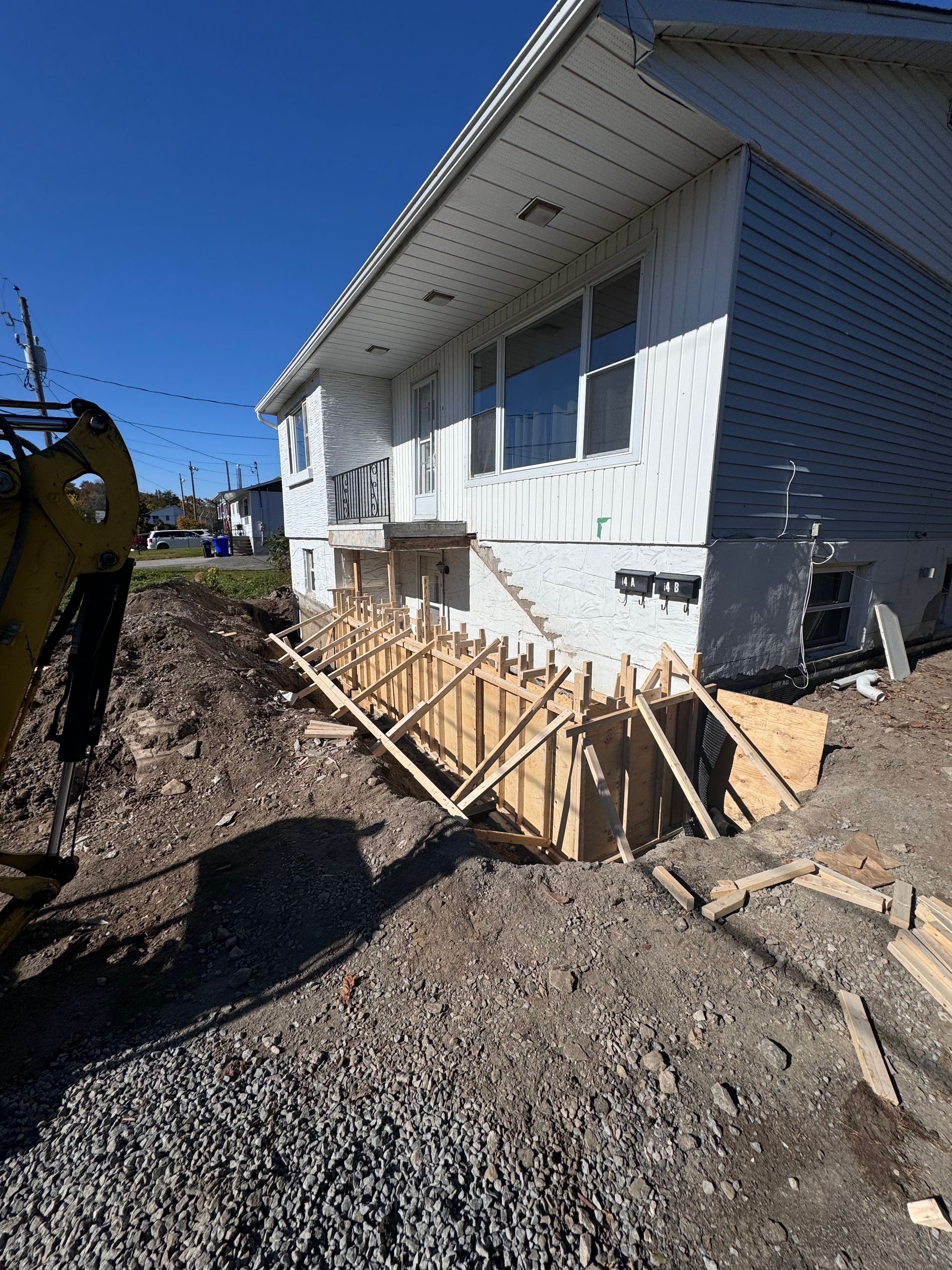 Extérieur d'une maison blanche avec des travaux de réparation des fondations, une tranchée de gravier et des supports en bois sous un ciel d'un bleu éclatant
