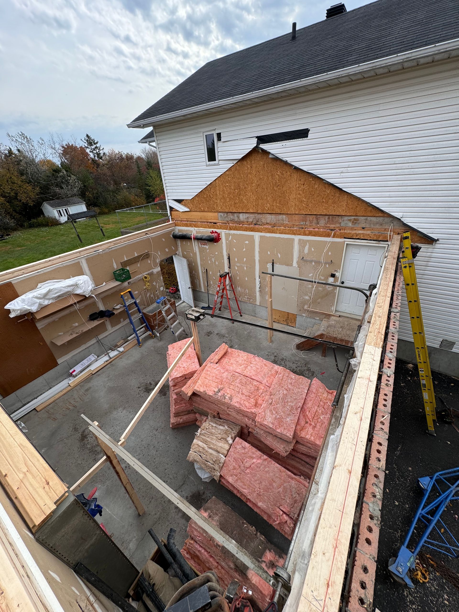 Chantier de construction dans une cour arrière, avec des murs à ossature bois, de l'isolant rose et une maison blanche en arrière-plan.