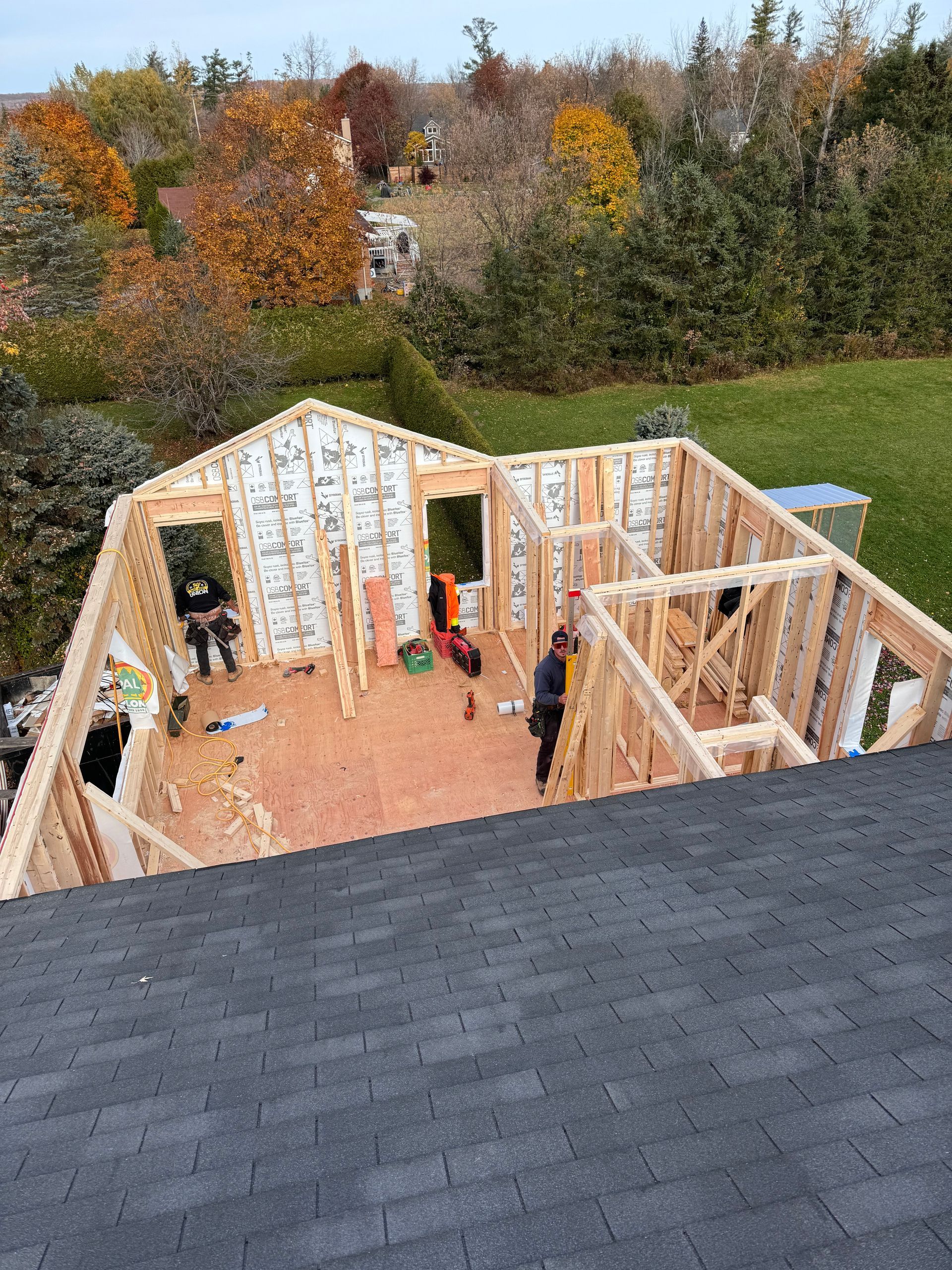 Maison en construction partielle avec charpente apparente et sous-plancher orange, vue d'un toit au milieu d'arbres d'automne