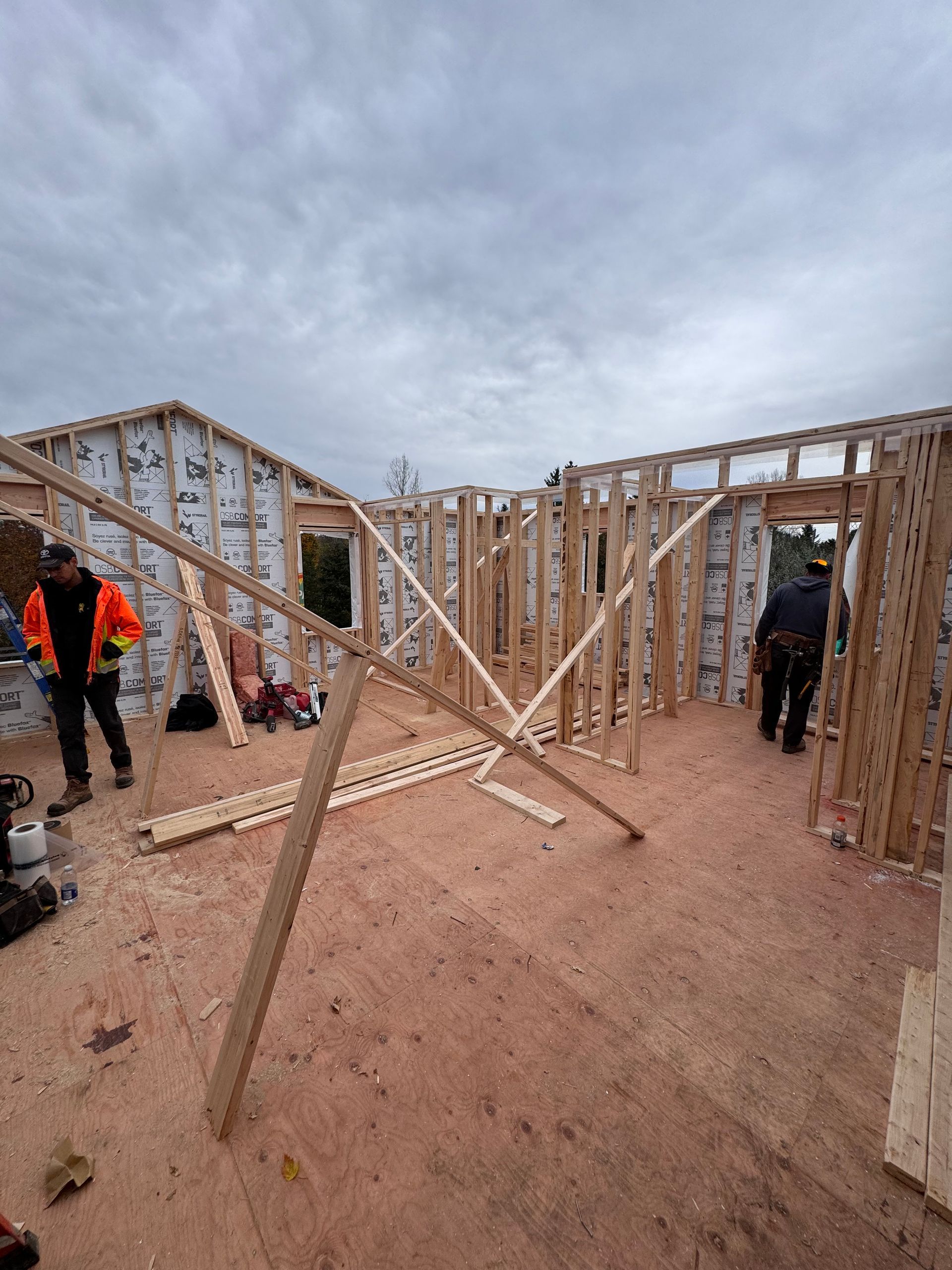 Charpente en bois d'une maison en construction sur un terrain vague sous un ciel nuageux, avec des ouvriers à proximité.