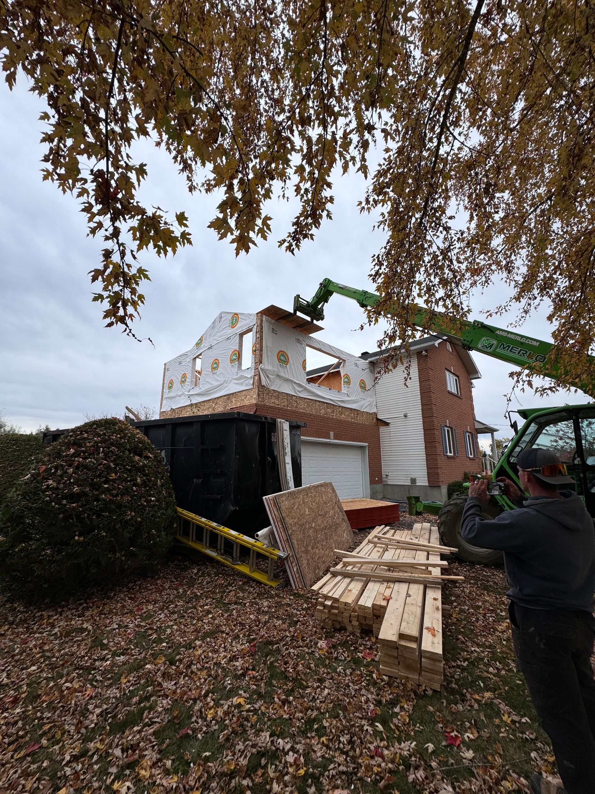 Chantier de construction avec un bâtiment en briques en rénovation, nacelle élévatrice verte et palettes en bois dans une cour recouverte de feuilles.