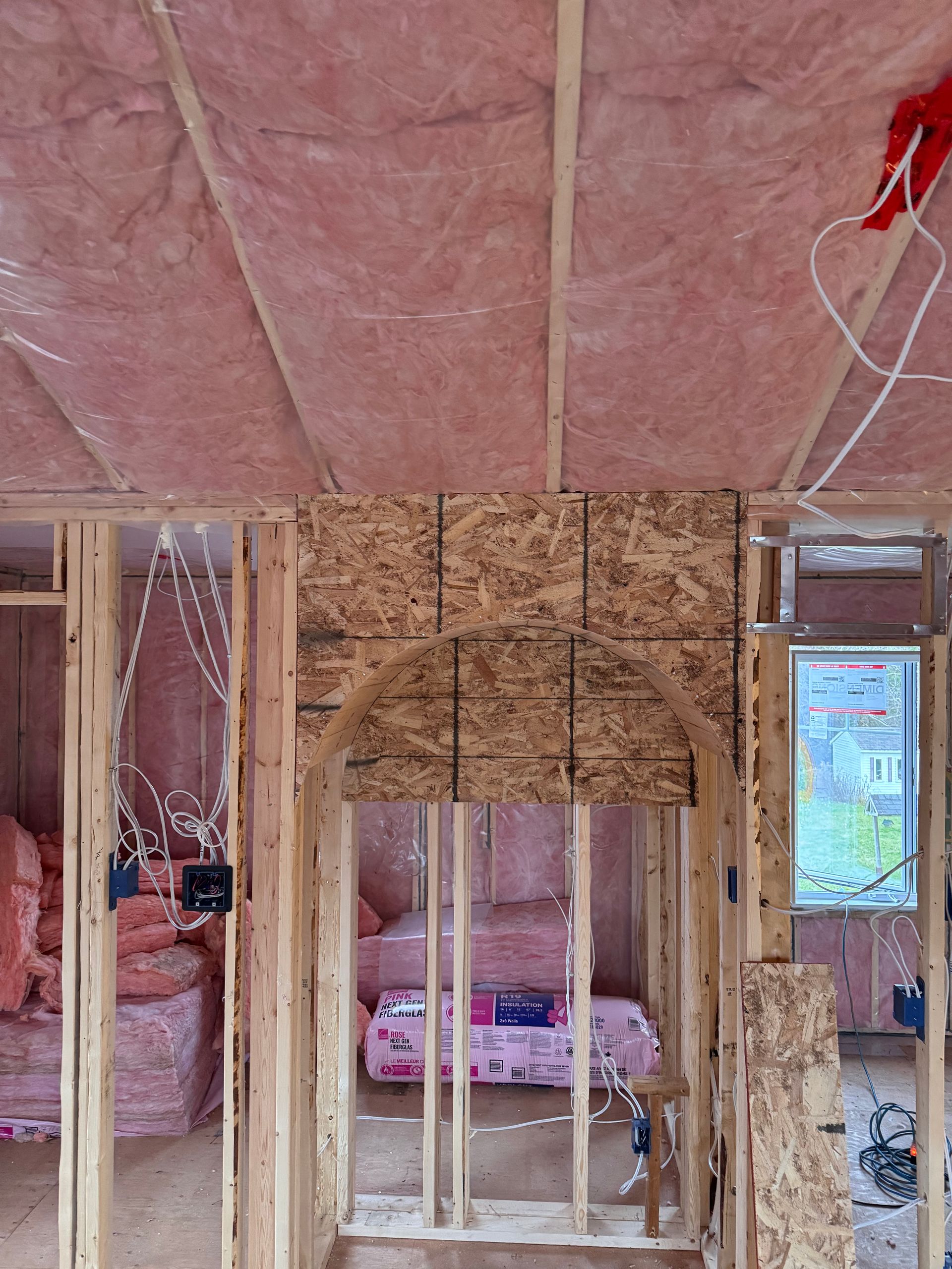 Pièce partiellement ossature en construction avec isolation apparente, mousse de plafond rose et une porte donnant sur une chambre.