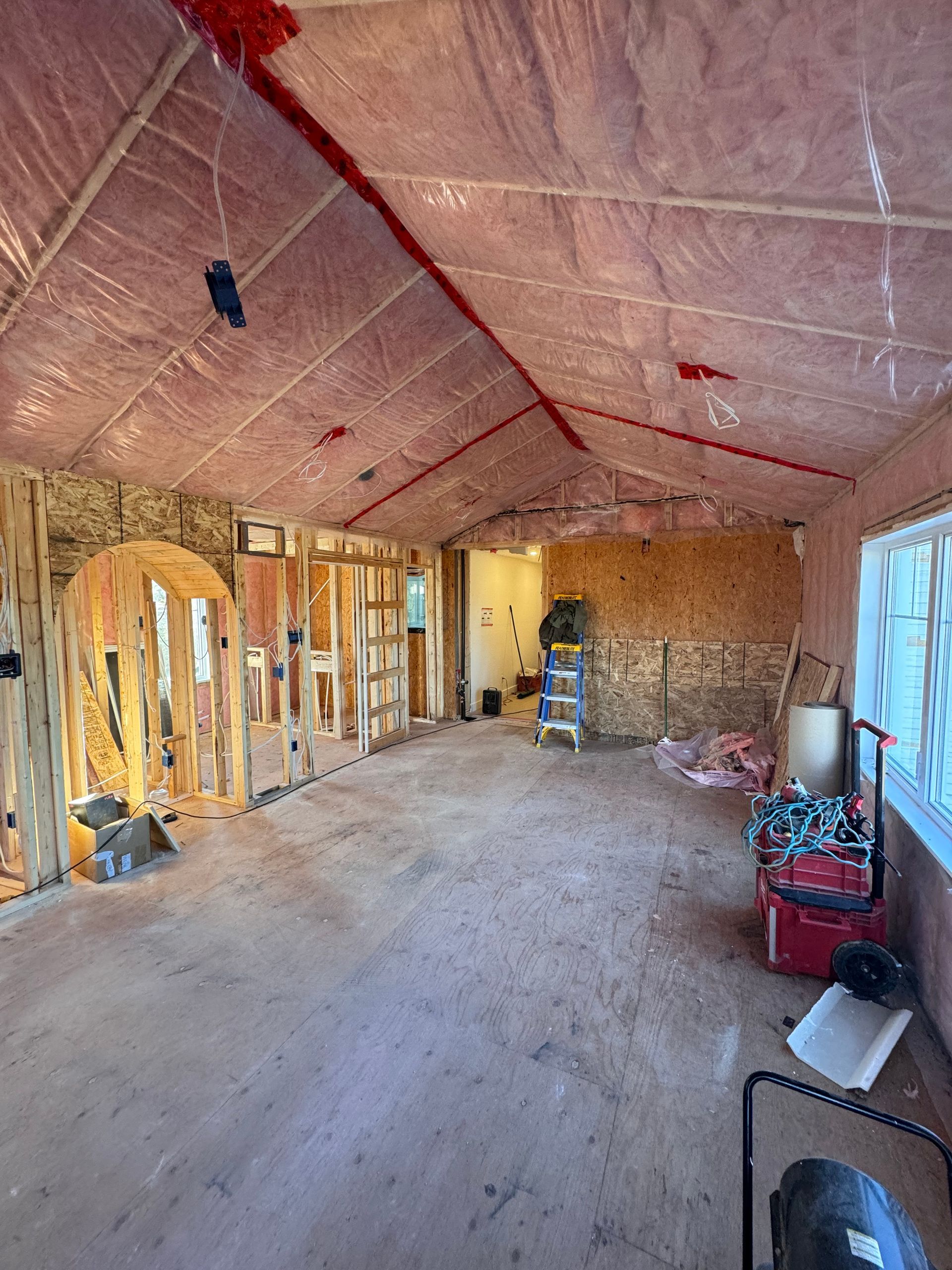 Grenier non aménagé avec isolation apparente, plancher en contreplaqué et outils près d'une fenêtre et de murs à ossature.