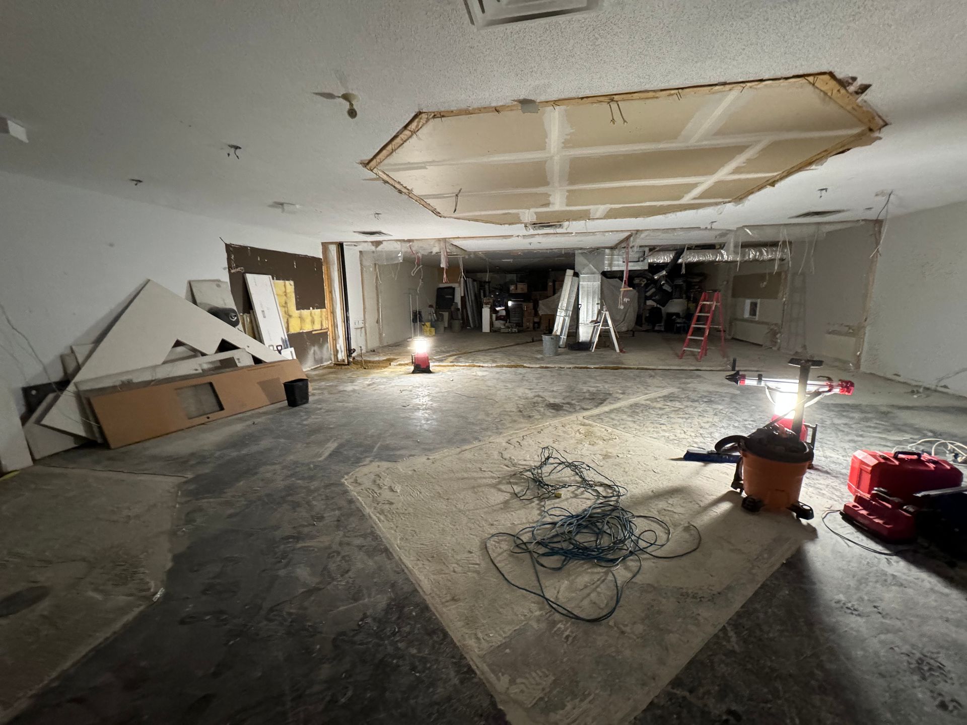 Travaux de rénovation en cours dans une pièce endommagée, avec plafond apparent, débris et lampes de travail au sol.