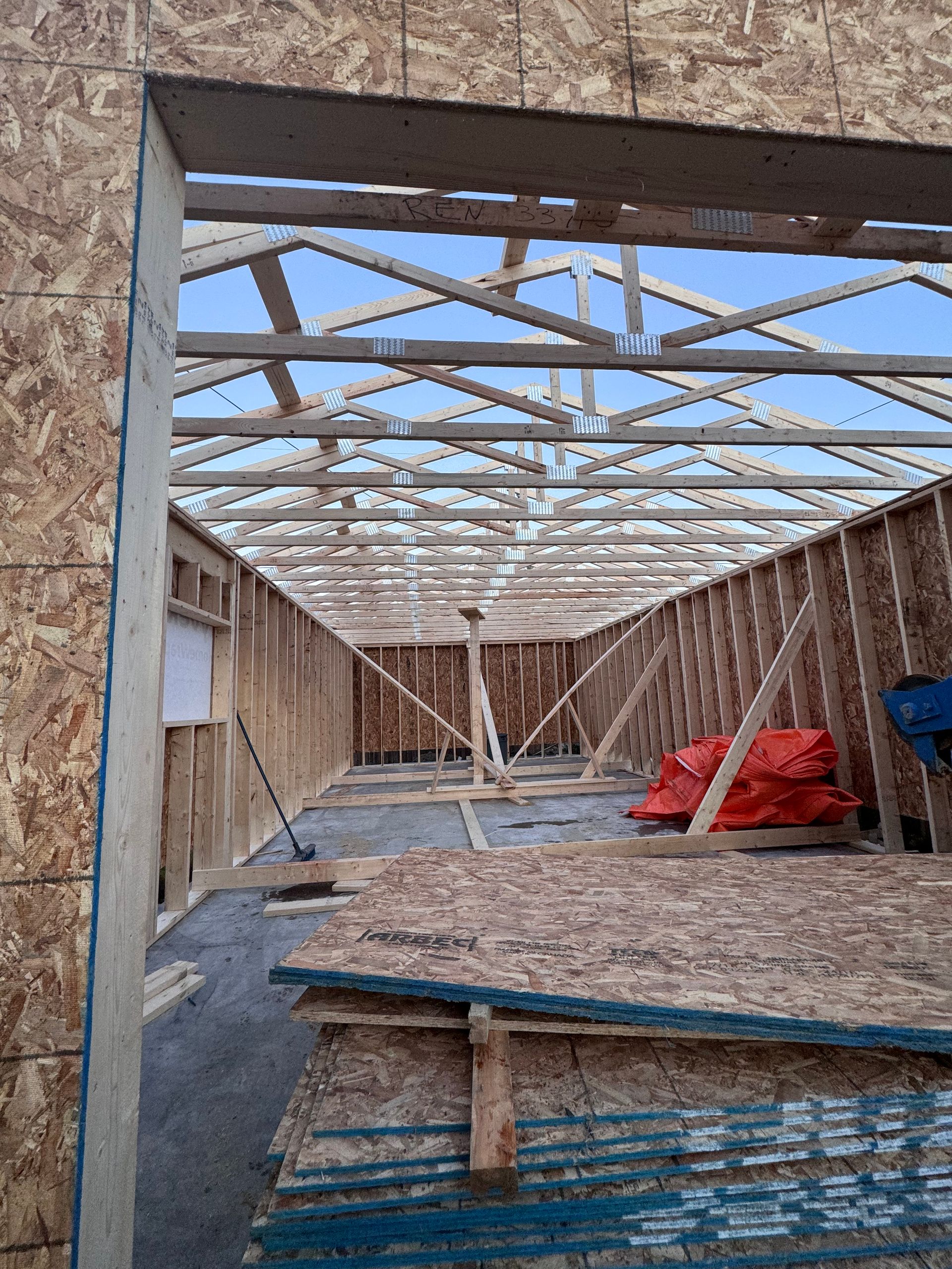 Intérieur d'une maison en bois partiellement construite, avec charpente apparente, piles de contreplaqué et bâches rouges.