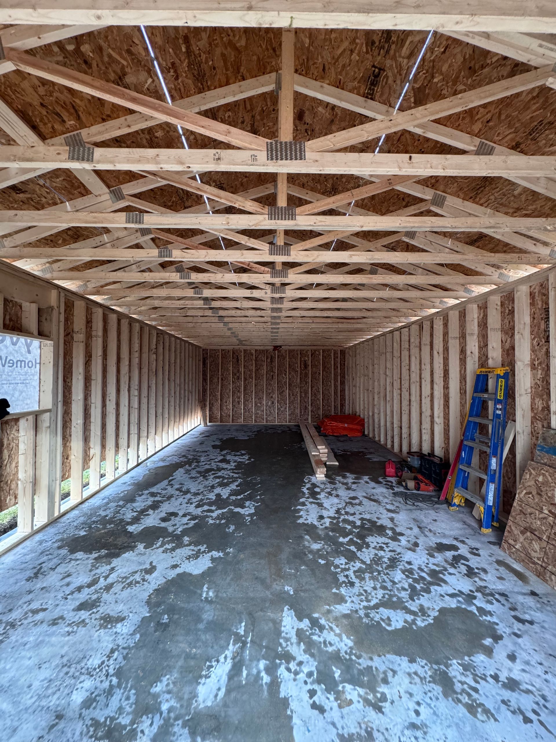 Intérieur d'un garage inachevé avec charpente en bois apparente, sol en béton et outils empilés sur la droite.