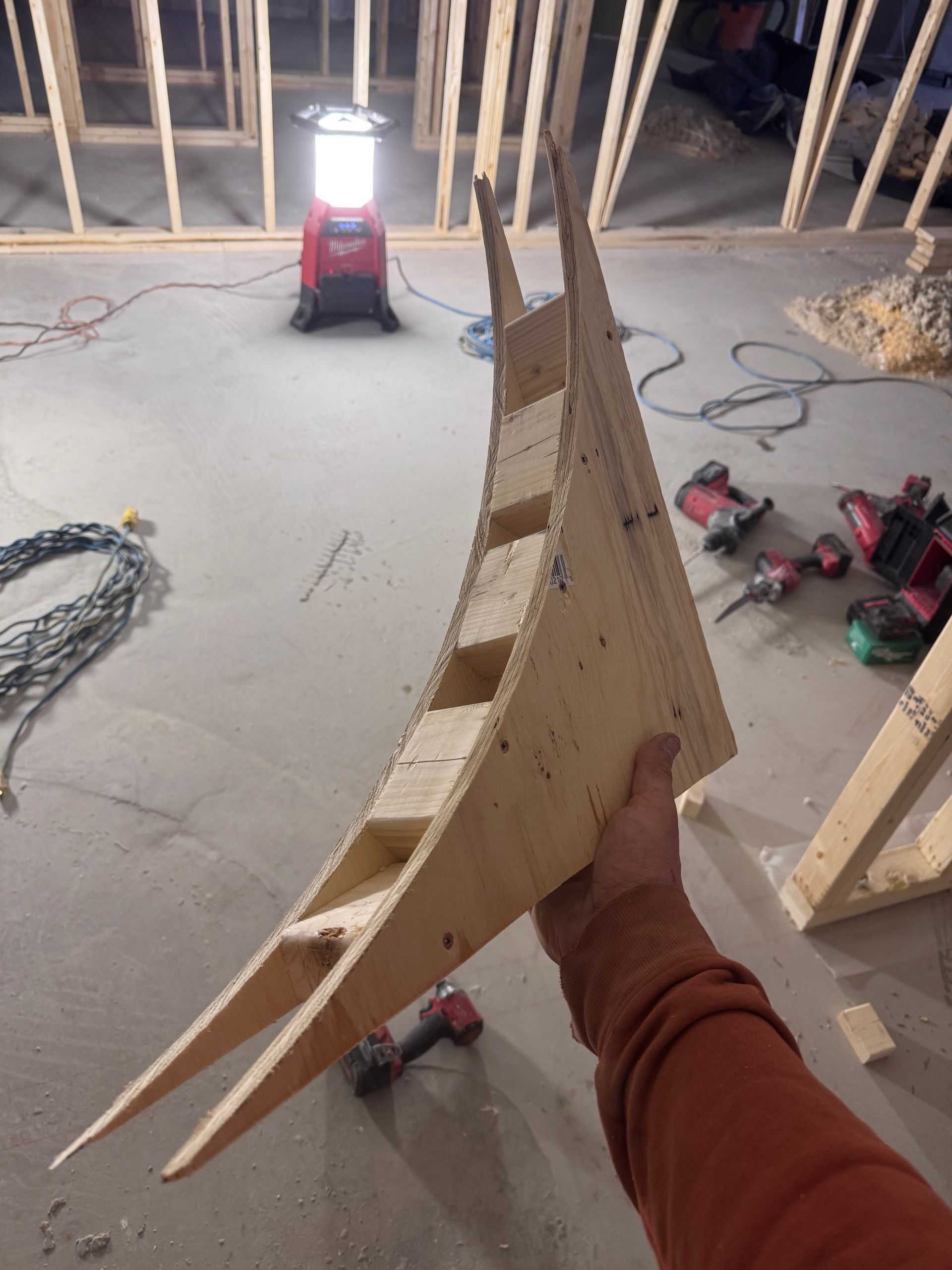 Une main tenant une planche de bois courbée dans une pièce inachevée avec des outils, une charpente apparente et une lampe de travail.