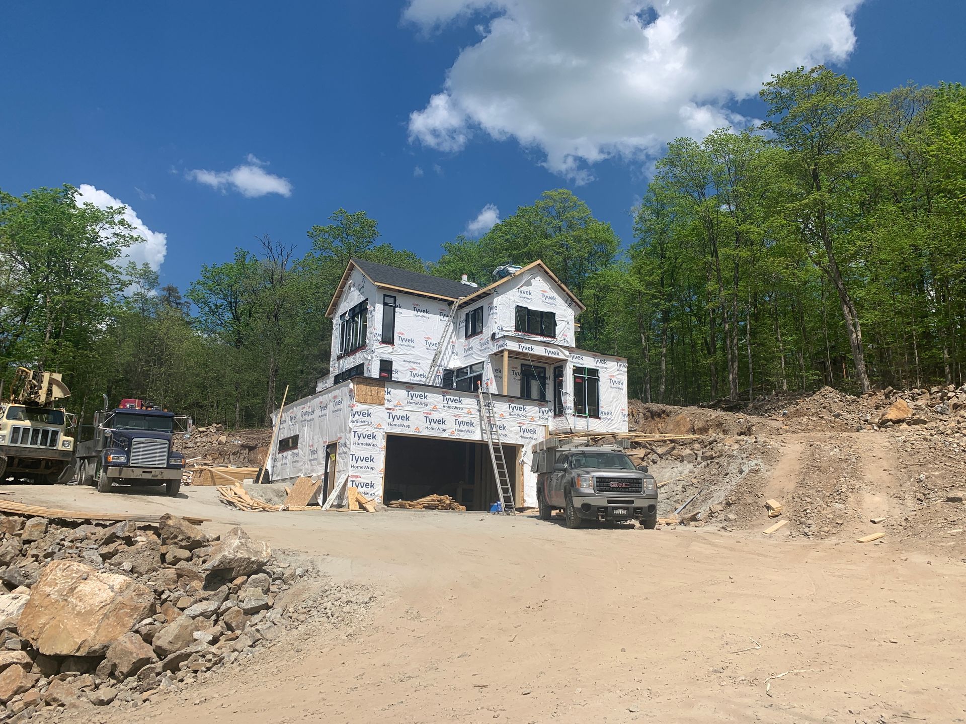 Maison blanche en construction sur un terrain vague avec deux camions et des arbres sous un ciel bleu