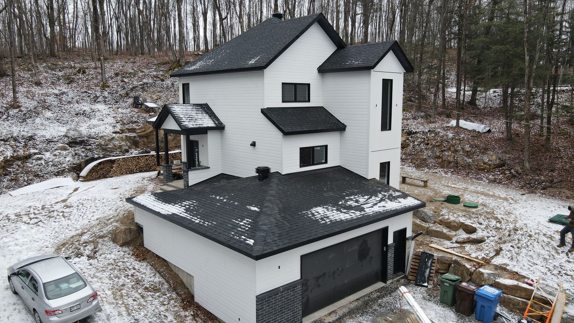 Maison à deux étages saupoudrée de neige, avec un toit noir, un garage et une voiture garée à côté, dans une cour boisée.