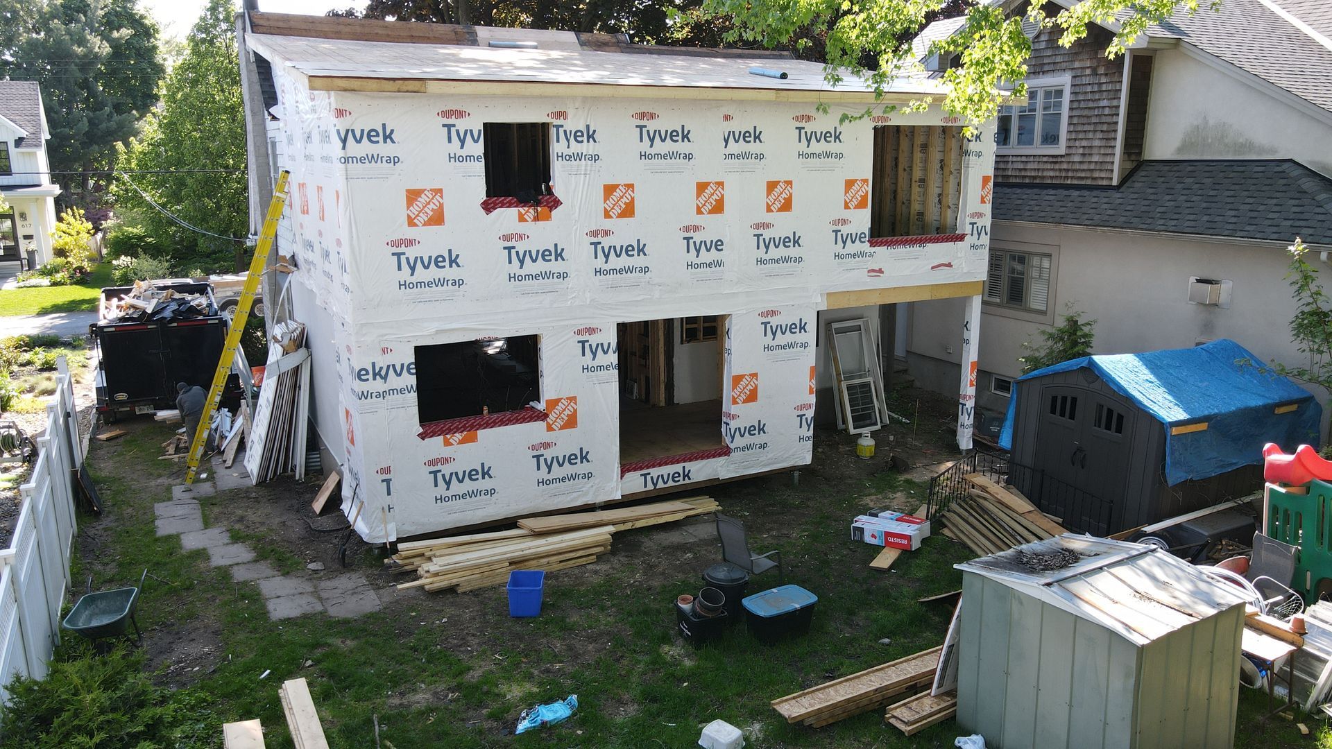 Maison en construction, enveloppée de Tyvek, fenêtres ouvertes et cour encombrée de matériaux.
