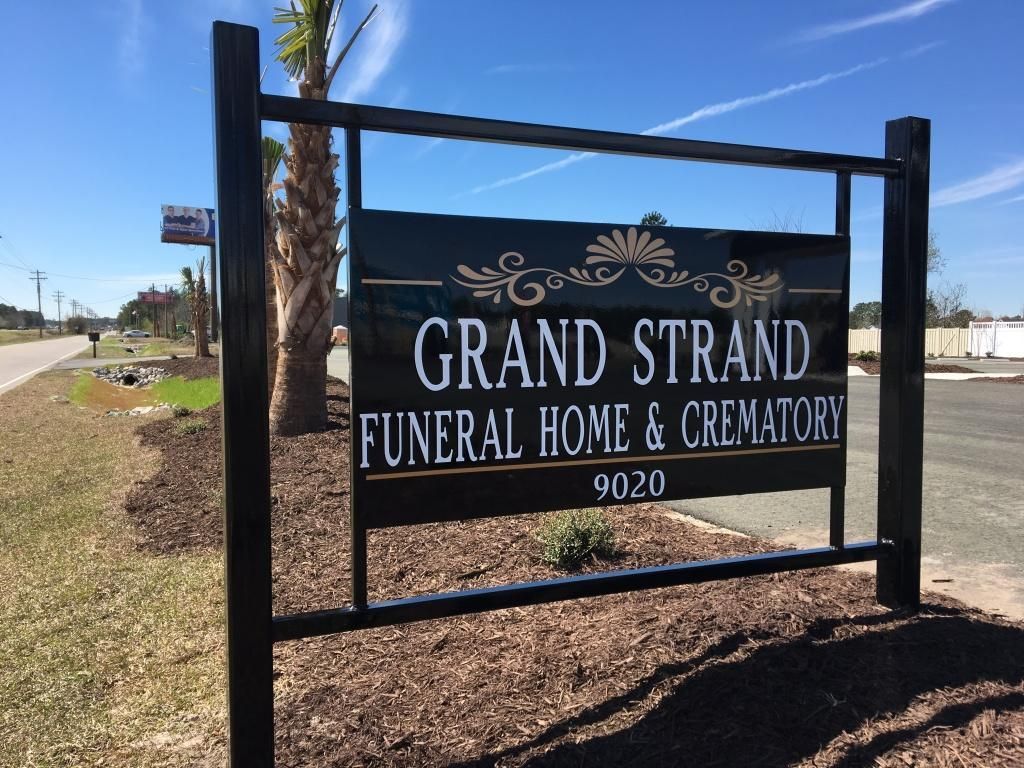 A black outdoor sign for Grand Strand Funeral Home & Crematory at 9020, situated near palms on a sunny day.