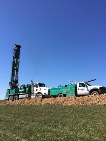 Two utility trucks and a drilling rig on a grassy field under a clear blue sky