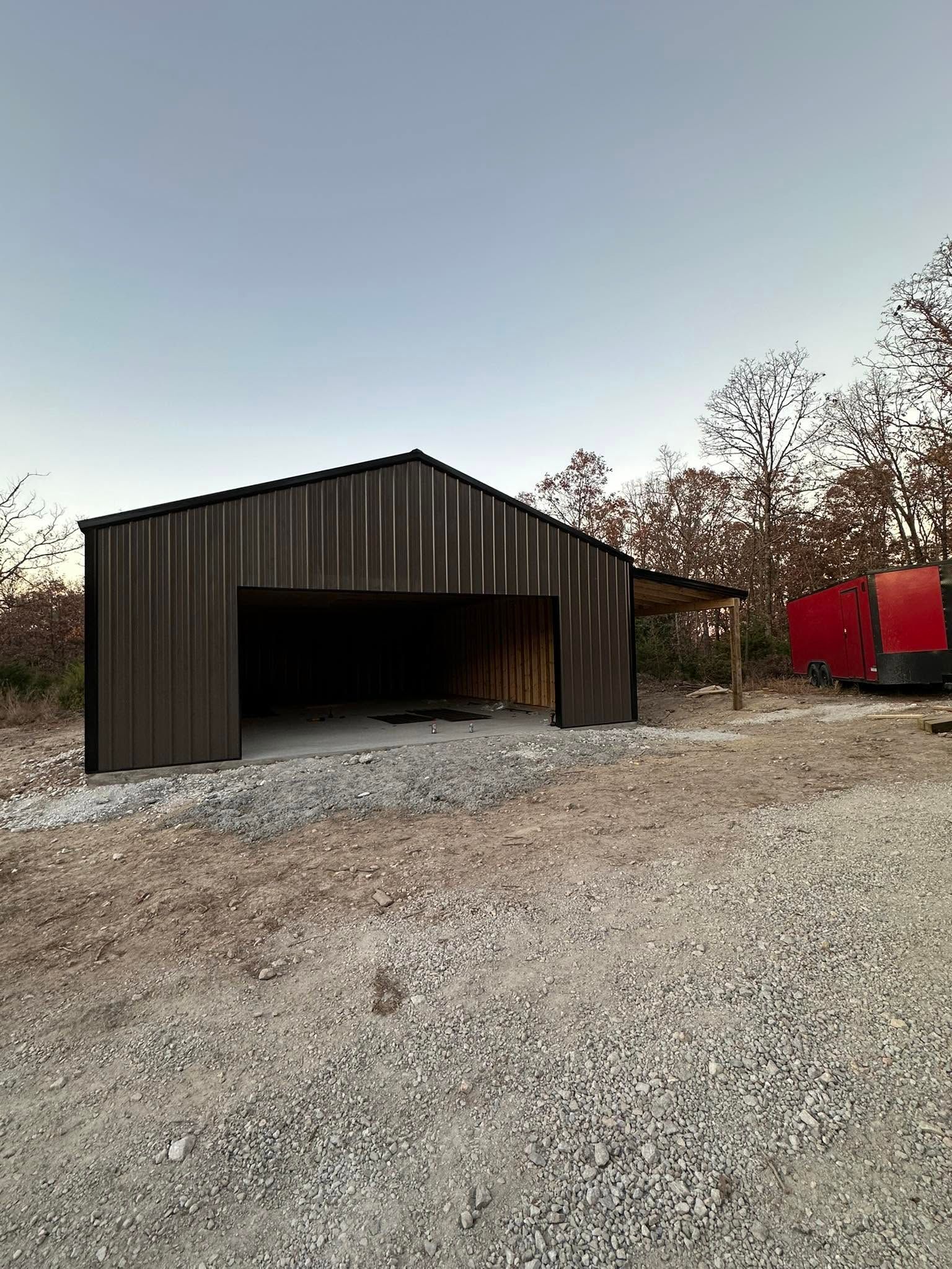 Dark wooden barn on gravel lot with red trailer and leafless trees at dusk