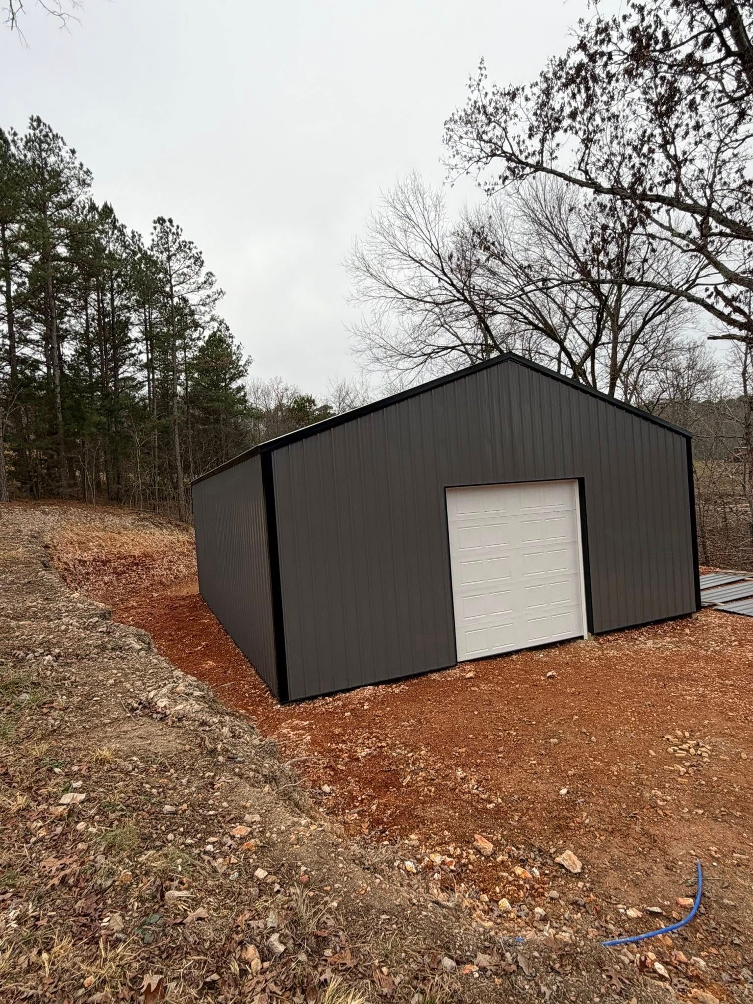 Gray metal shed with white door on a wooded hillside covered in brown leaves.