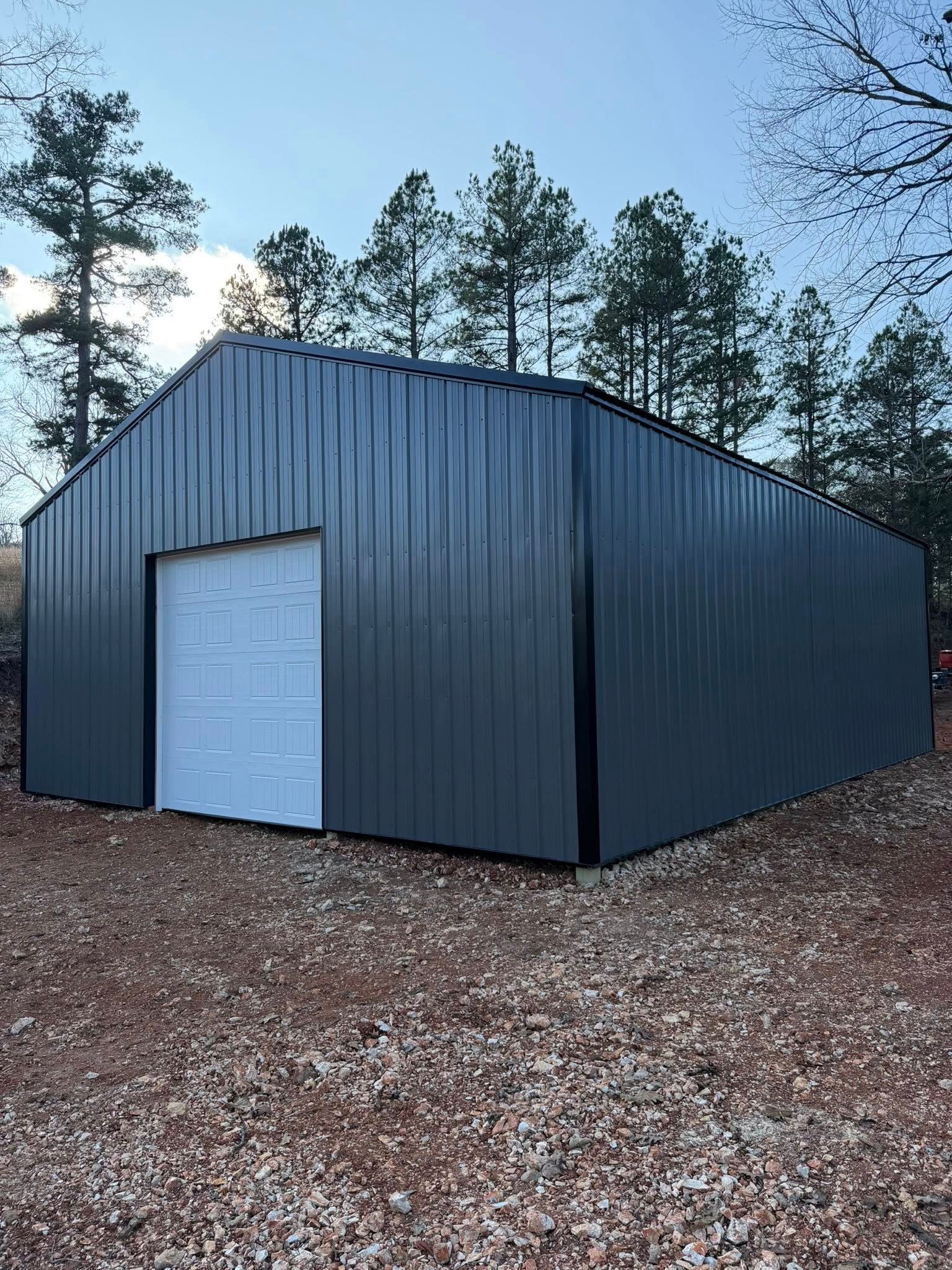 Dark gray metal shed with a white door on a gravel lot, surrounded by pine trees.