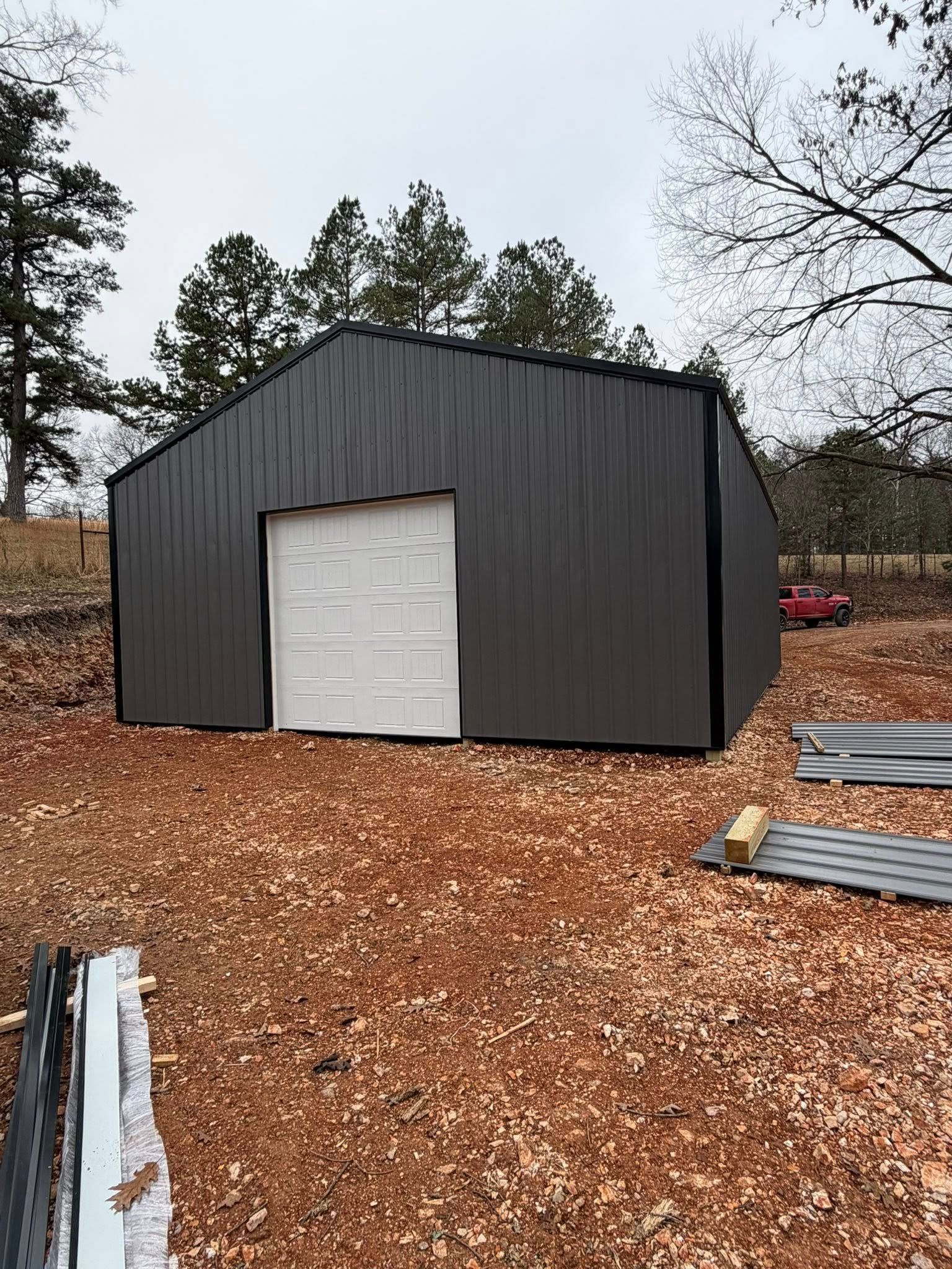 Black metal shed with a white garage door on a woodchip lot, surrounded by trees