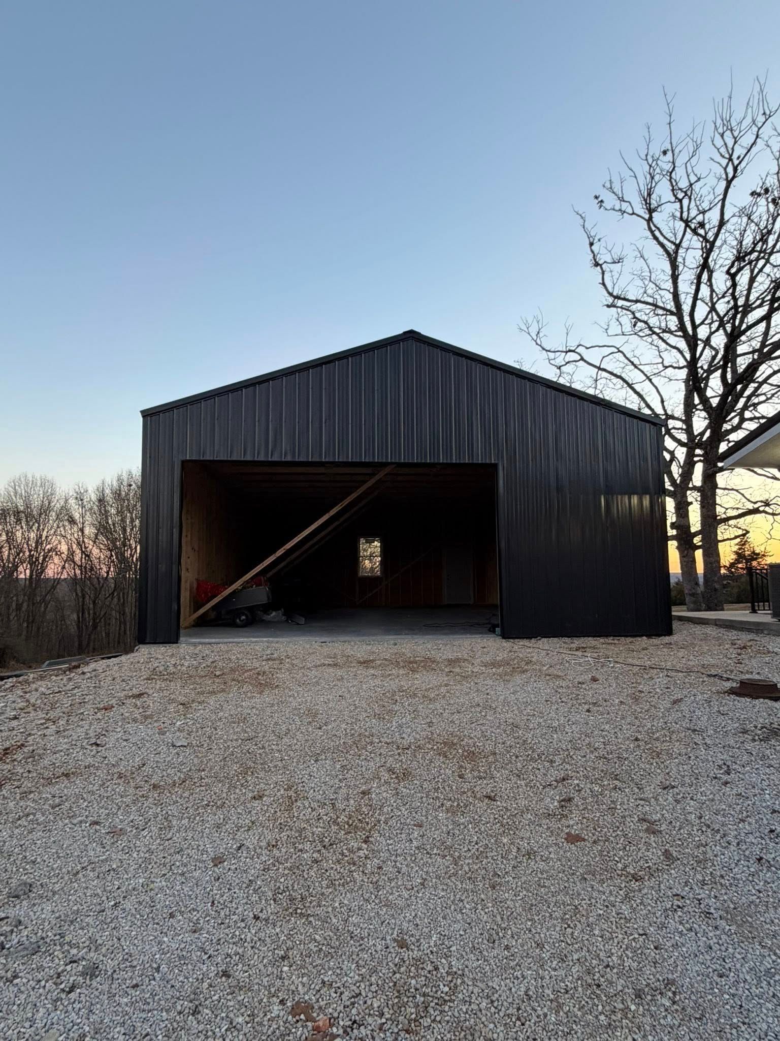 Black barn with open front on a gravel lot at dusk, with bare trees and a pale sky.