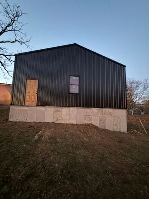 Black metal-clad building on a concrete foundation at dusk, with a door, window, and bare tree branches nearby