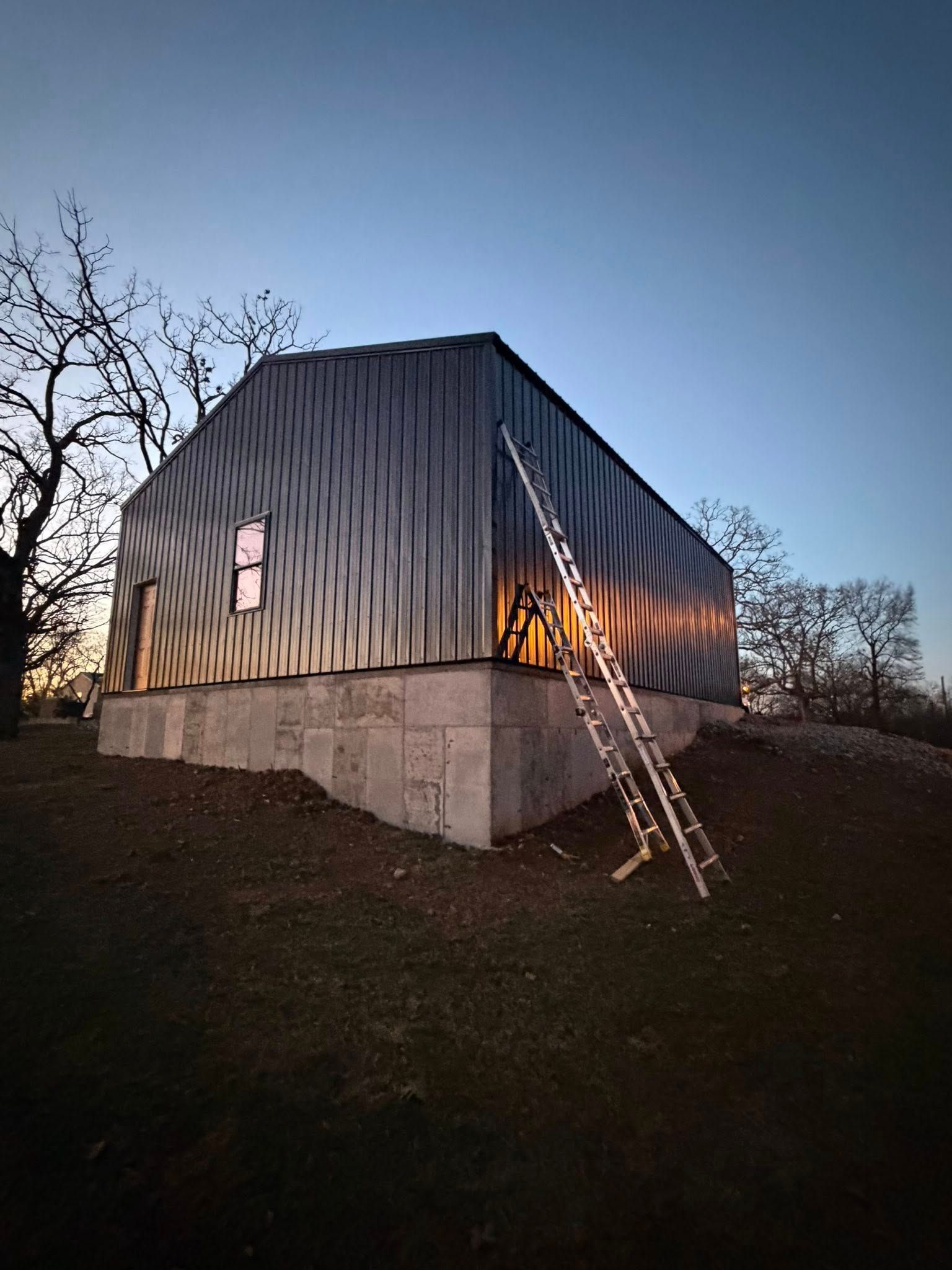 White metal-clad house on a concrete base at dusk, with a ladder leaning against the side.