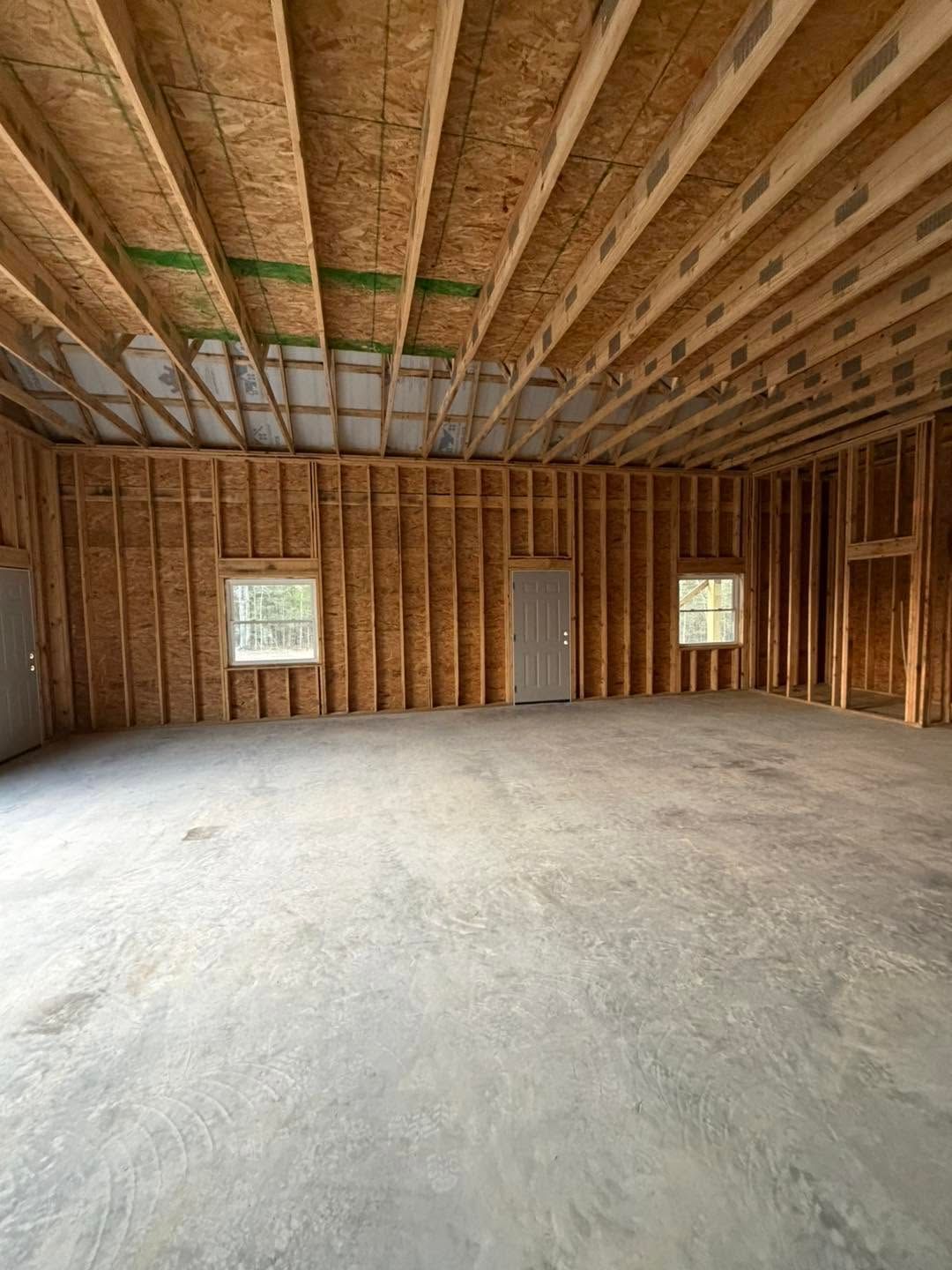 Empty unfinished room with exposed wood framing, concrete floor, and two windows and a door.