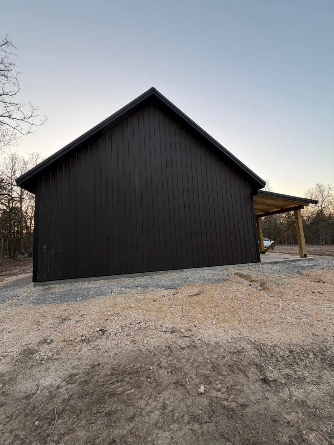 Black wooden barn with a gravel foreground and a small covered side entrance at dusk