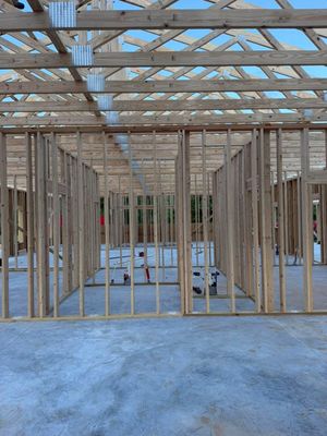 Wood-framed house under construction, showing interior studs and roof trusses on a concrete slab.