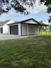 White metal garage with dark doors beside a gravel drive, under trees beside a grassy yard.
