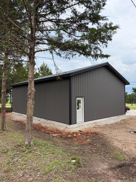 Dark gray metal shed with a white door, beside trees on a dirt lot.