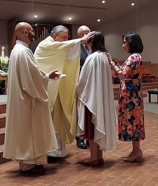 A person in a white robe stands in prayer, hands pressed together, holding a rosary with a cross.