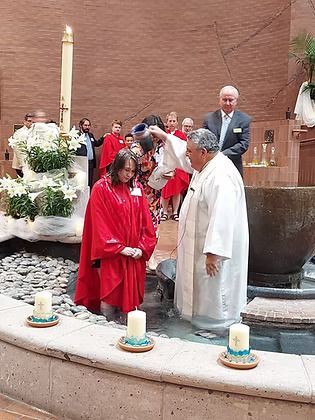 A person in a red robe kneels in a church baptismal font, being baptized by a clergyman in white.