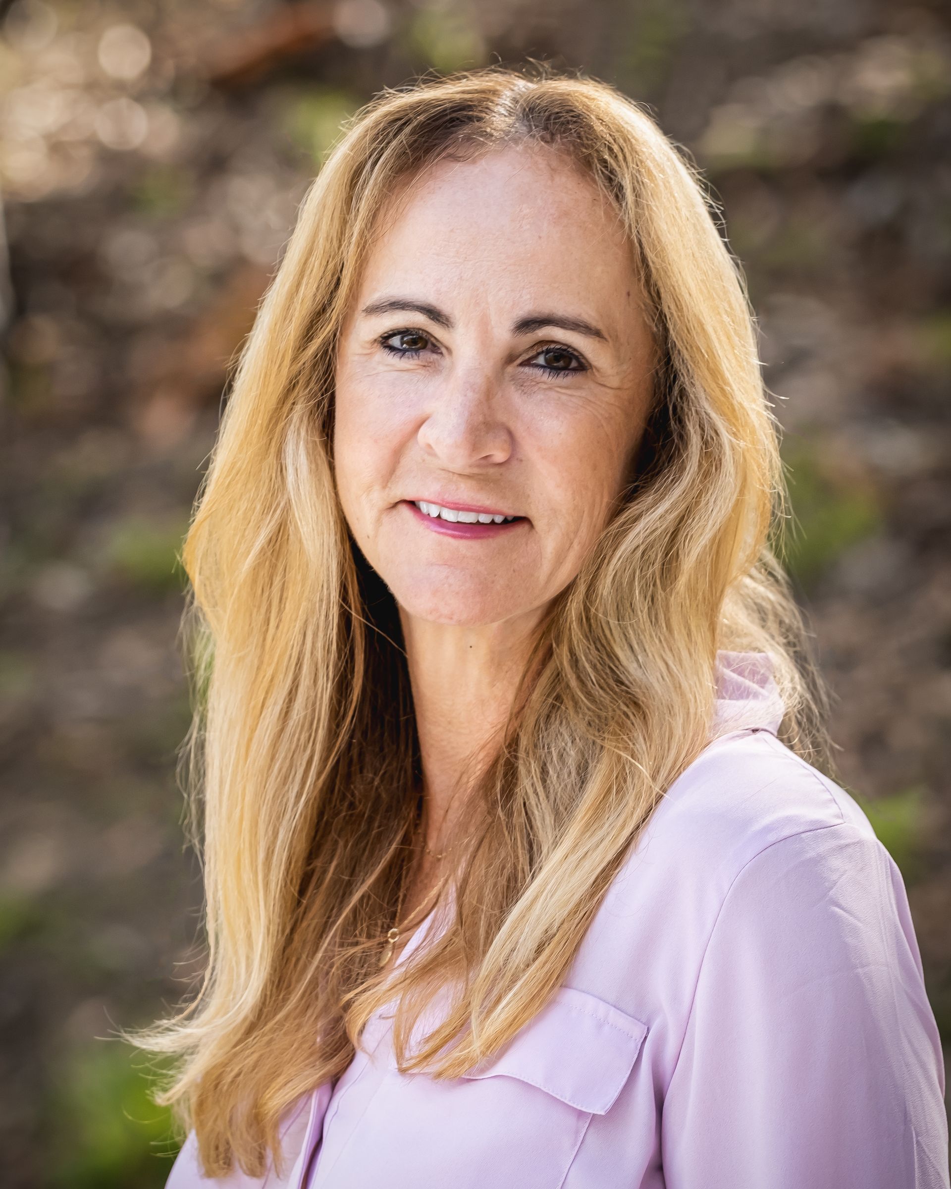 A woman with long blonde hair is wearing a pink shirt and smiling for the camera.