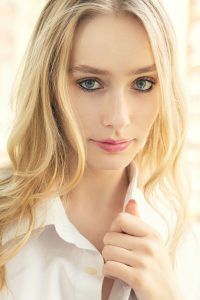A woman with long blonde hair and blue eyes is wearing a white shirt.