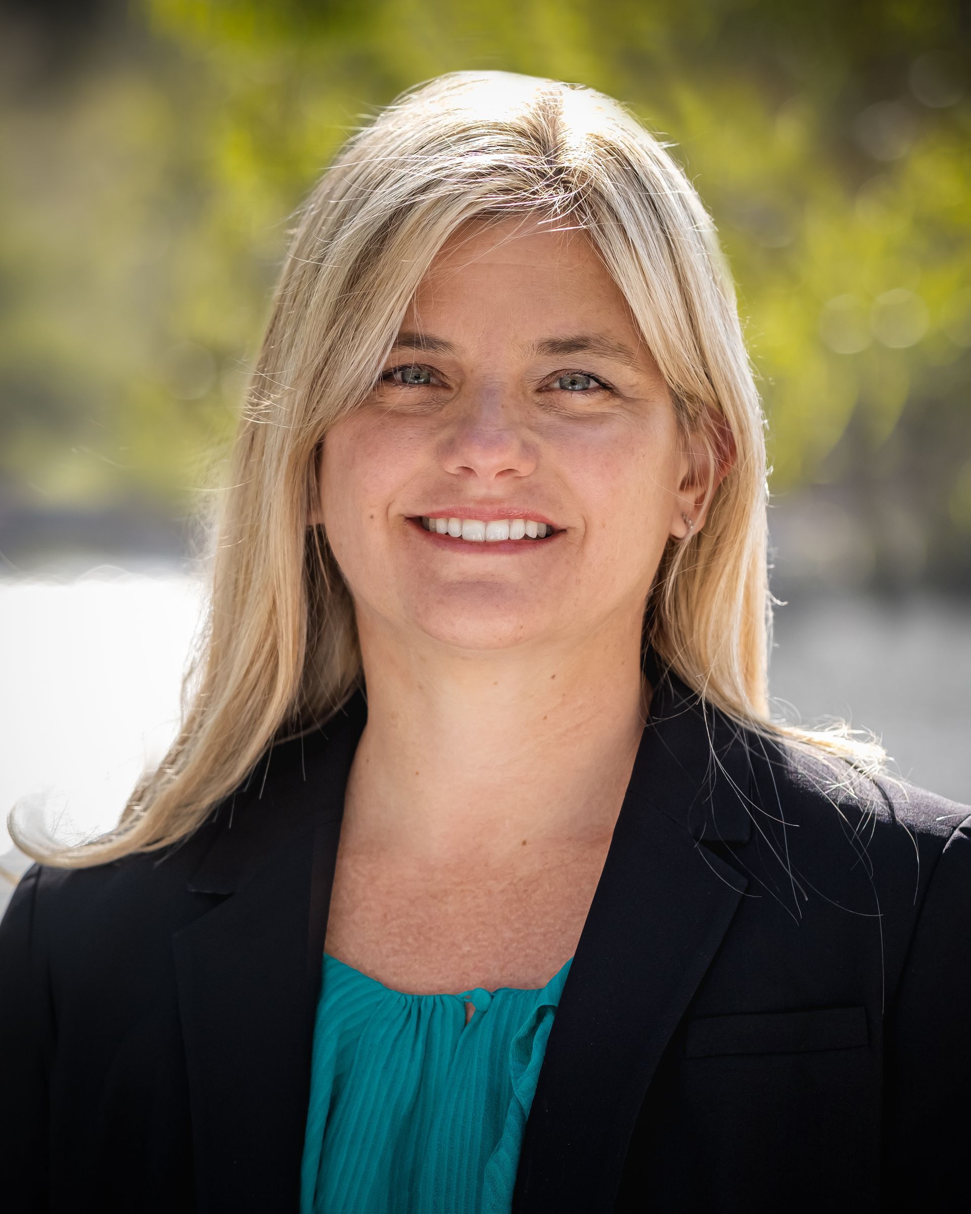 A woman in a blue shirt and black jacket is smiling for the camera.