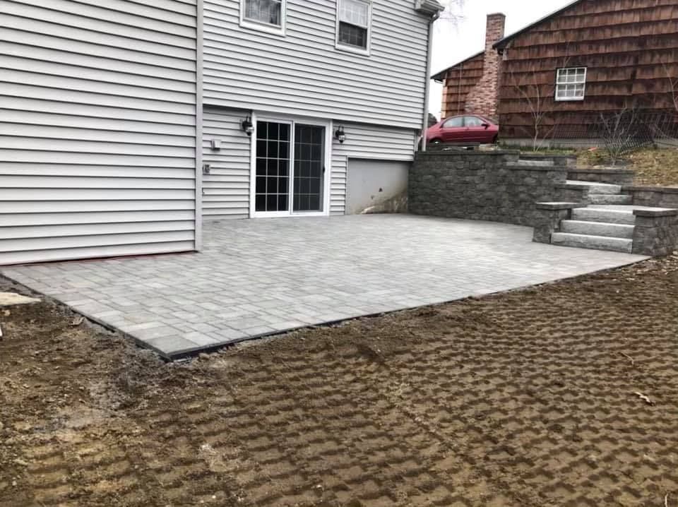 Paver patio beside a house, with retaining wall steps and a small yard area.