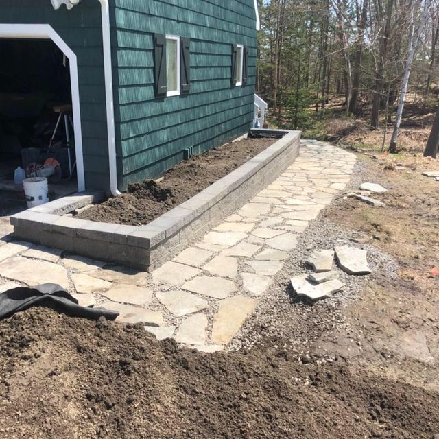 Stone walkway beside a green house under construction, with soil and retaining wall along the path