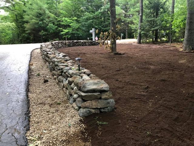 Stone-lined garden bed beside a paved road in a wooded area